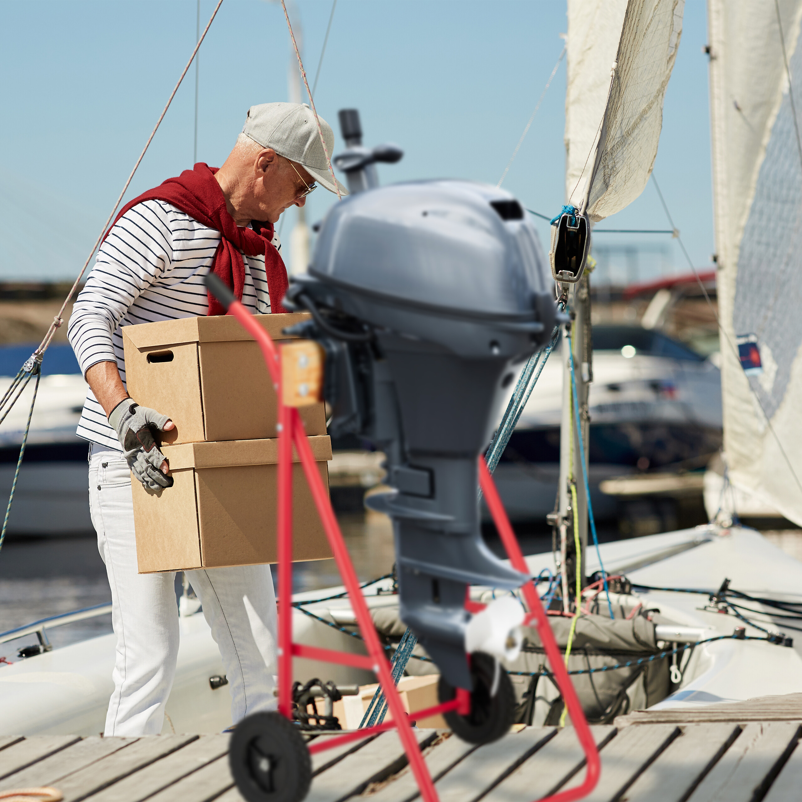 Älterer Mann in Kapitänsmütze und gestreiftem Shirt trägt Kartons auf Segelboot.