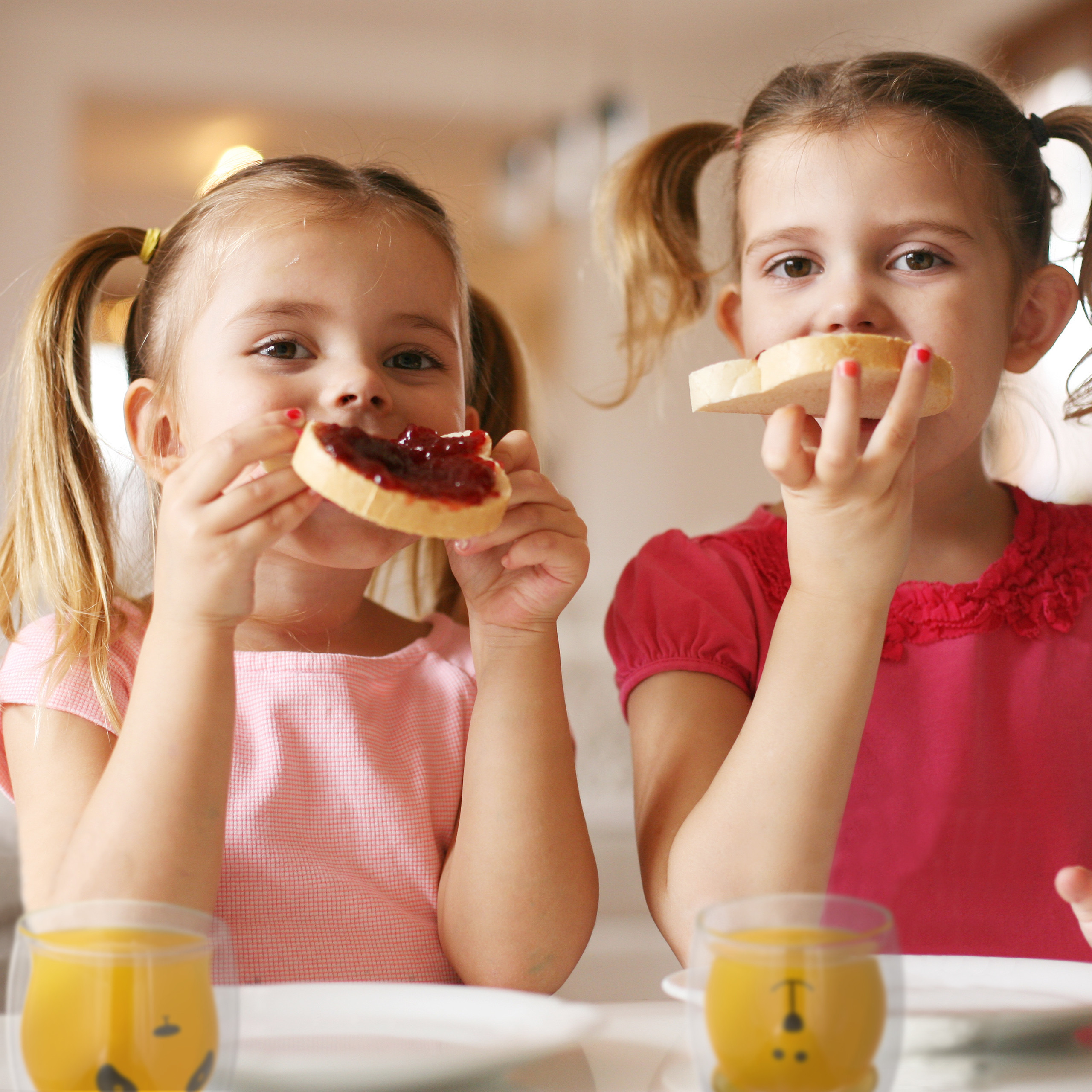 Zwei kleine Mädchen essen Brot mit Marmelade und trinken Orangensaft am Tisch.