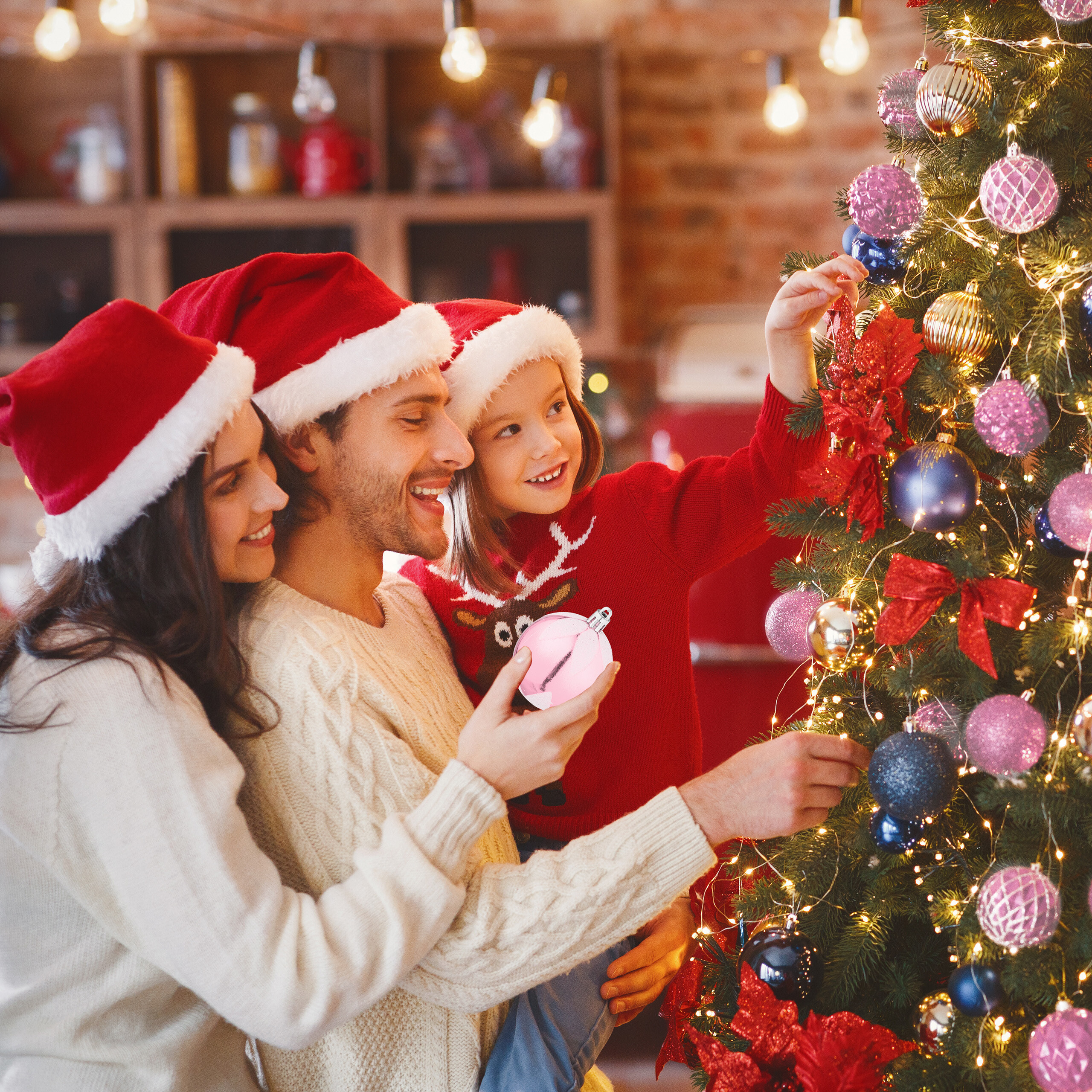 Eine Familie mit Weihnachtsmützen schmückt gemeinsam einen Weihnachtsbaum in einem gemütlichen Raum.