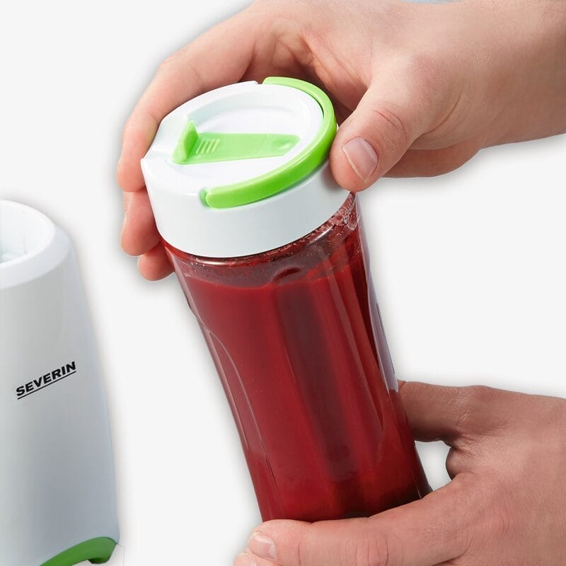 Manos enroscan una tapa blanca y verde en un vaso rojo junto a una base de licuadora.