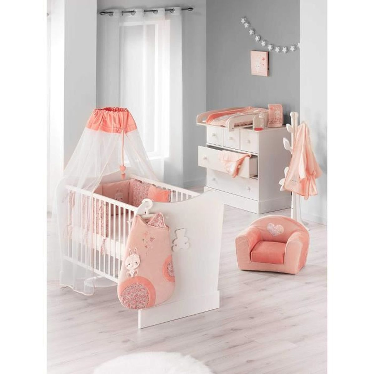 Ein Kinderzimmer mit weißem Gitterbett mit Himmel, Wickelkommode, einem kleinen rosa Sessel und Garderobe.