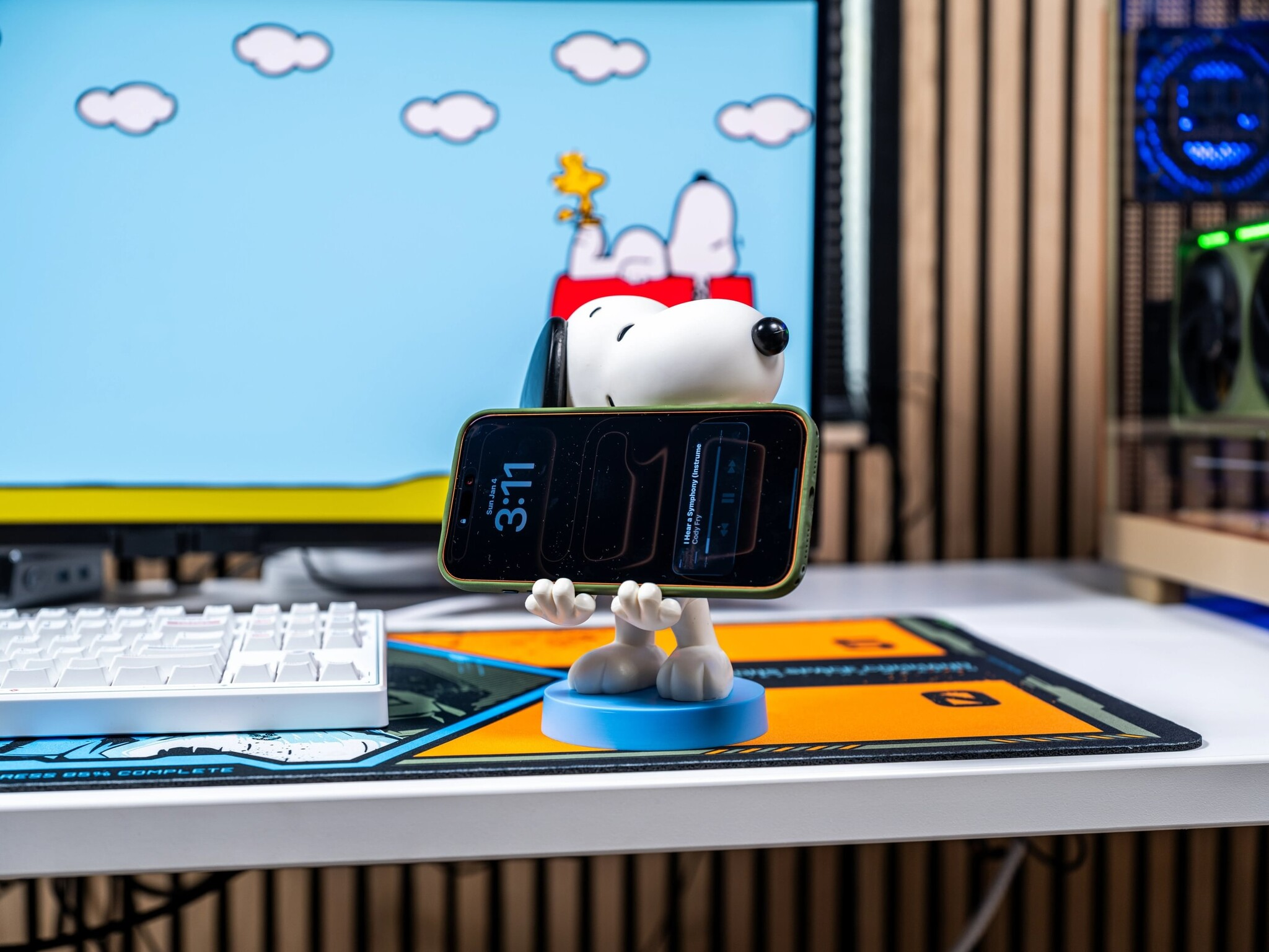 Snoopy-Figur hält ein Smartphone horizontal auf einem Schreibtisch vor einem Monitor mit Snoopy-Hintergrundbild.