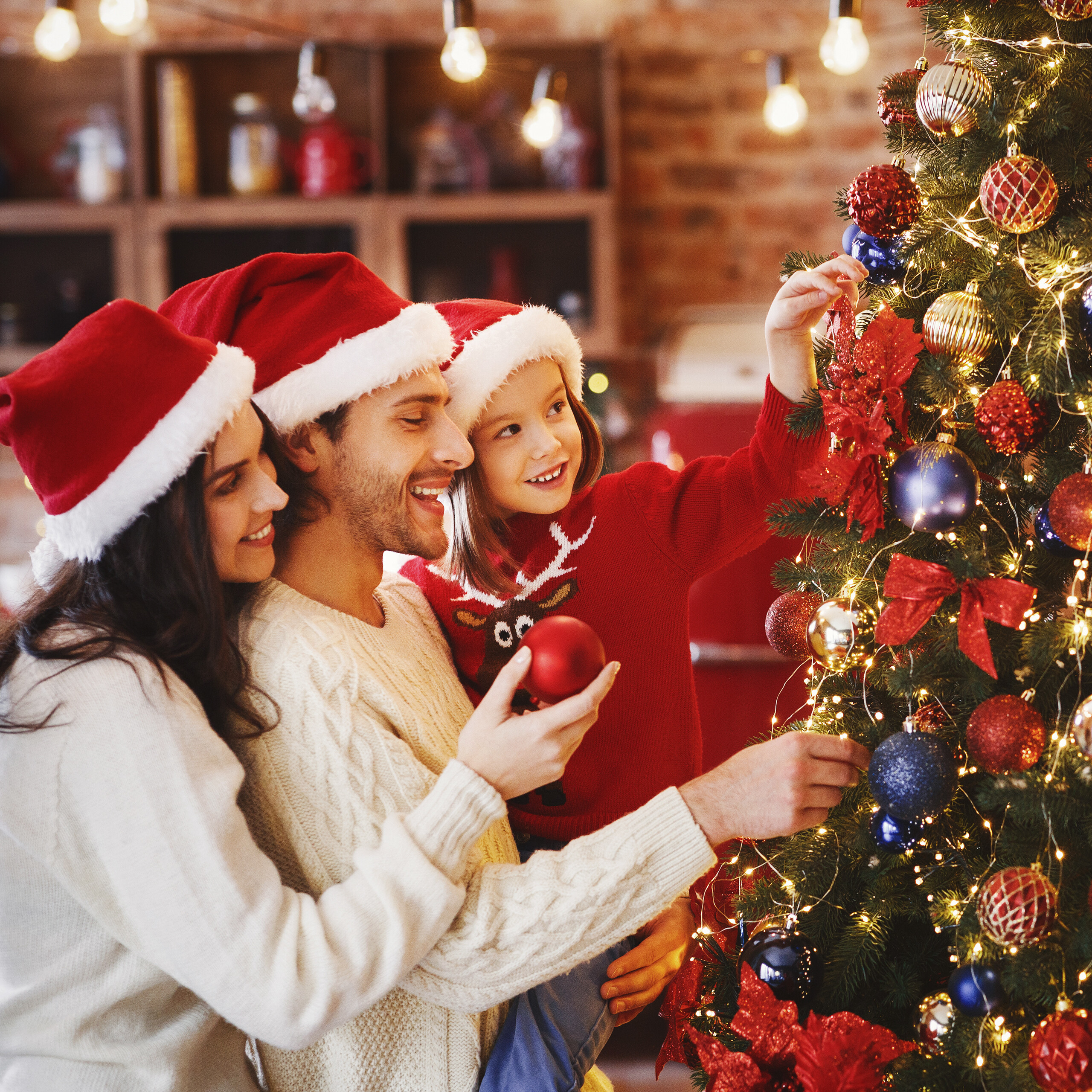Eine Familie mit Weihnachtsmützen schmückt gemeinsam einen Weihnachtsbaum in einem gemütlichen Raum.