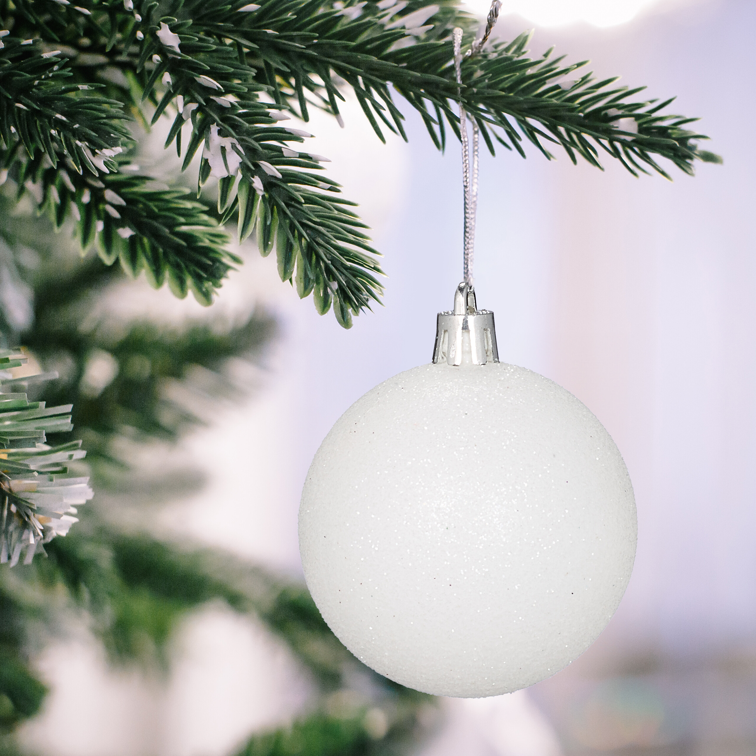 Eine glitzernde weiße Christbaumkugel hängt an einem grünen Tannenzweig vor verschwommenem Hintergrund.