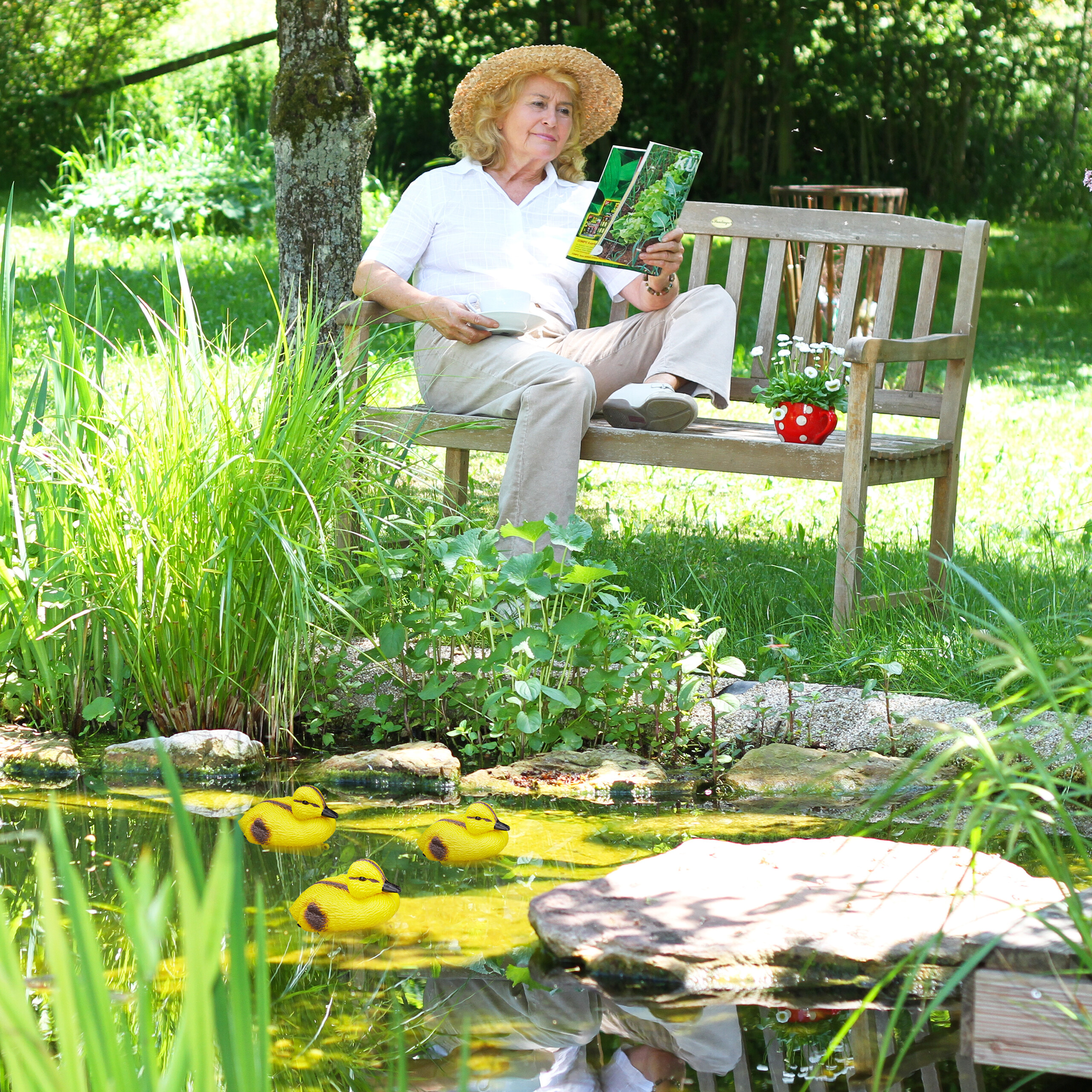 Eine blonde Frau mit Hut liest ein Buch auf einer Holzbank im Garten, nahe einem Teich mit kleinen Entenfiguren.