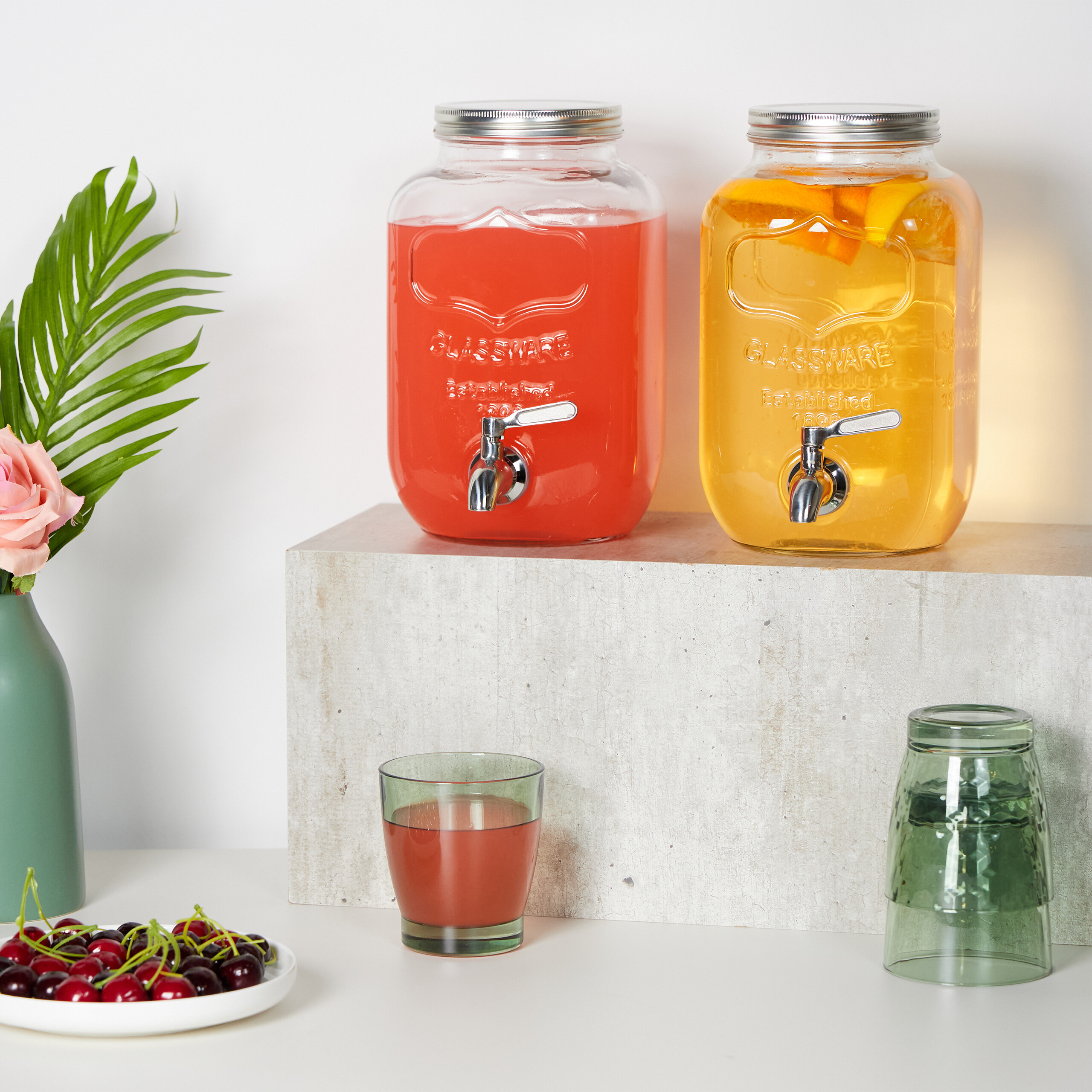 Zwei Glasbehälter mit rotem und orangenem Getränk auf einem Sockel, daneben Kirschen und Gläser.