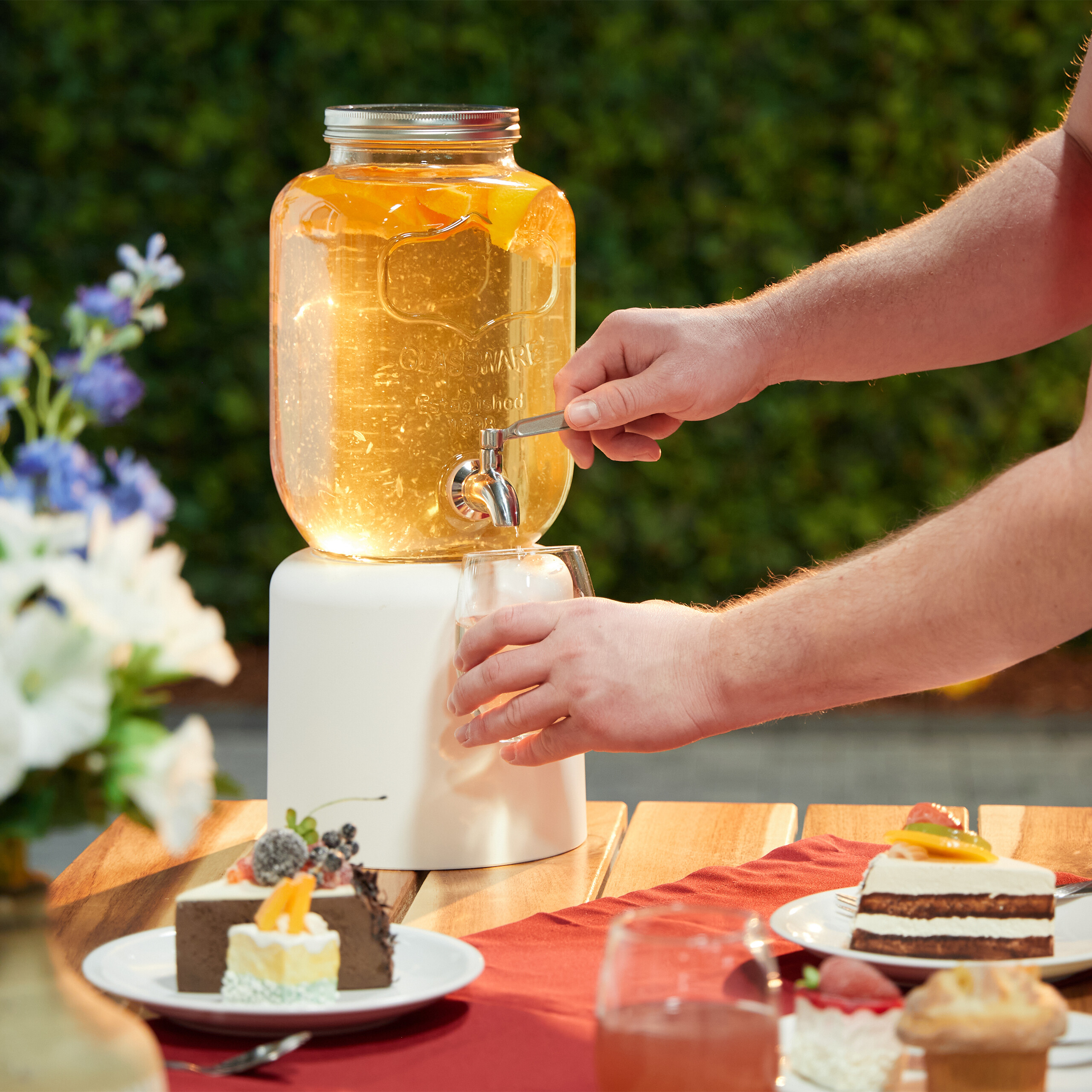 Eine Person gießt Getränk aus einem großen Glasbehälter mit Zapfhahn ein, auf Holztisch mit Kuchen.