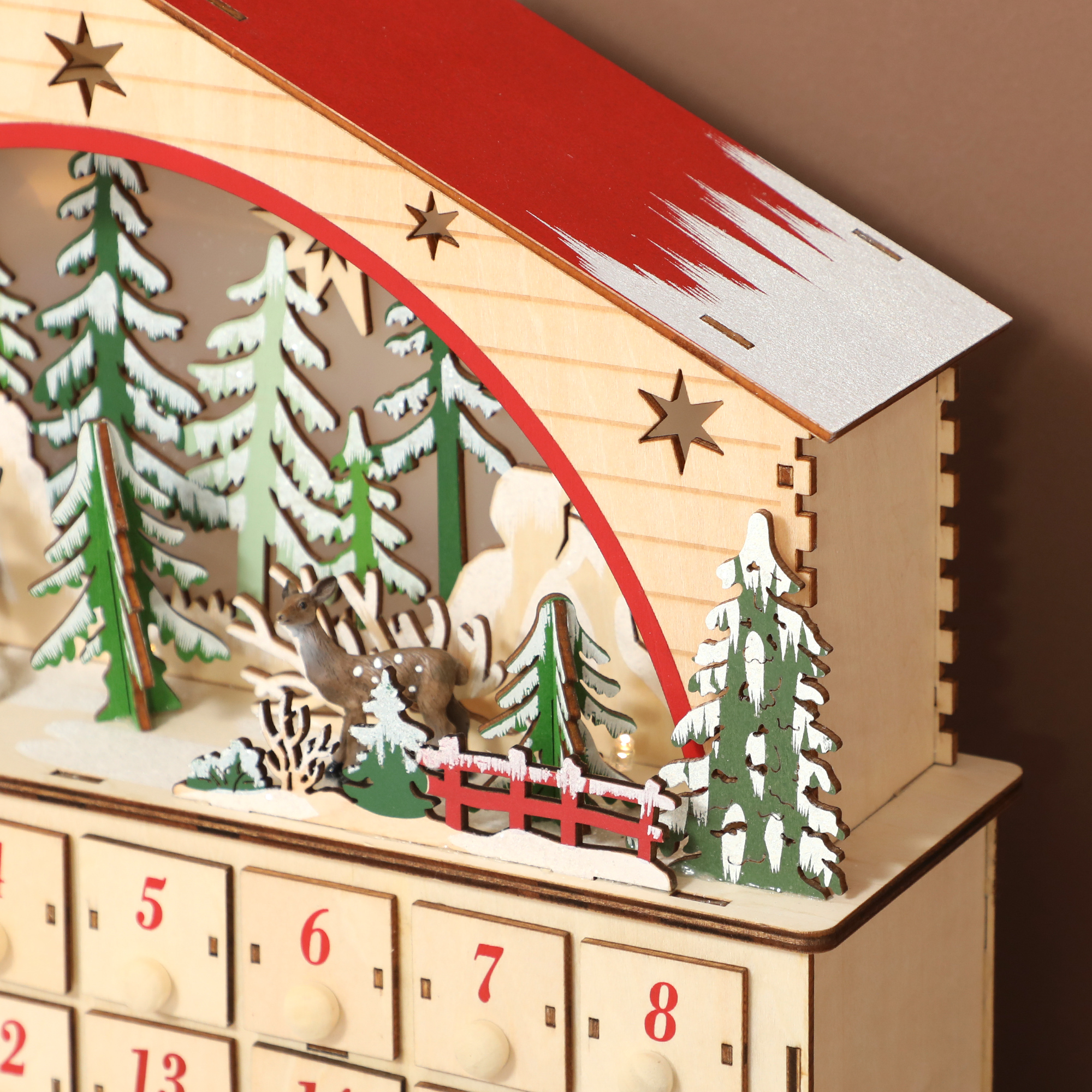 Calendario de adviento de madera con escena invernal, abetos y un cervatillo bajo un techo rojo.