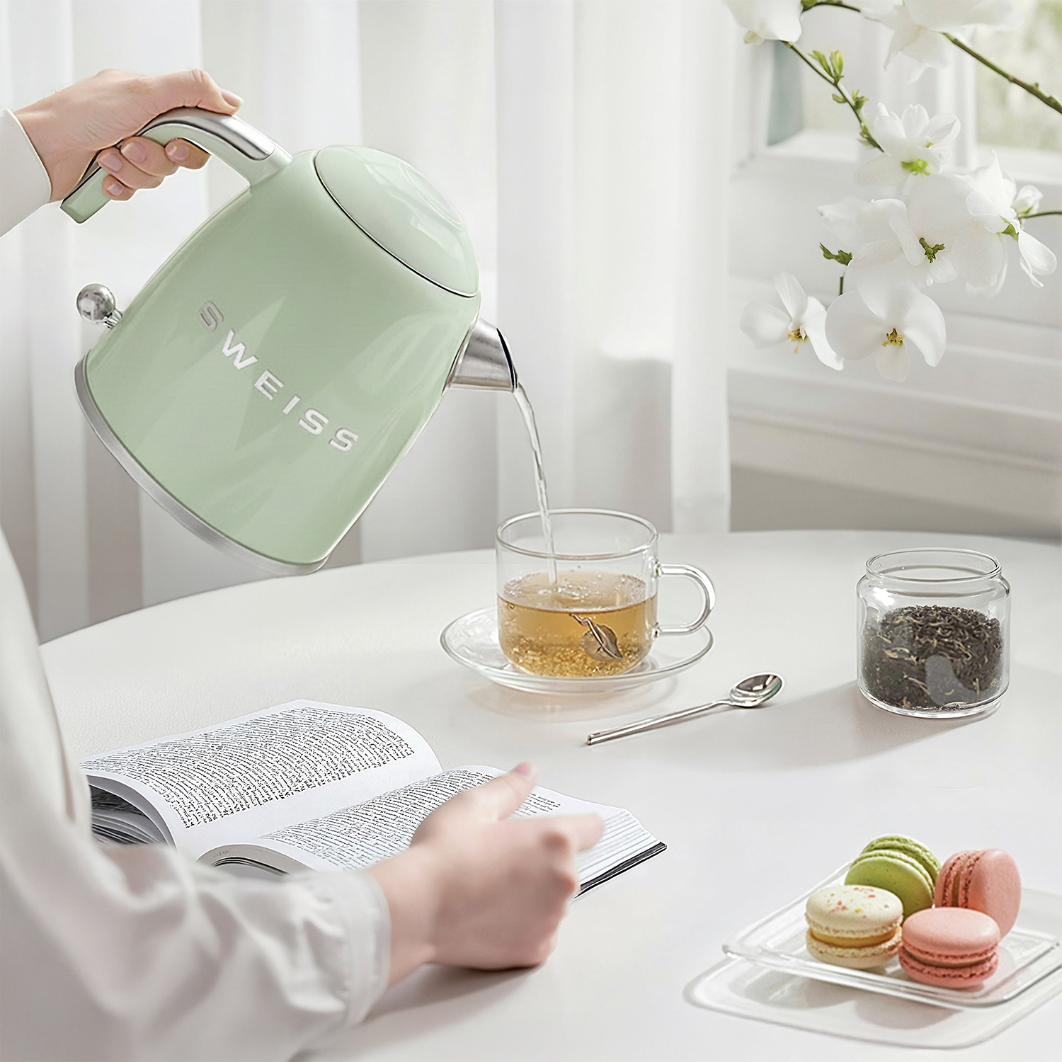Una mano vierte agua de un hervidor verde claro en una taza en una mesa con un libro y macarons.
