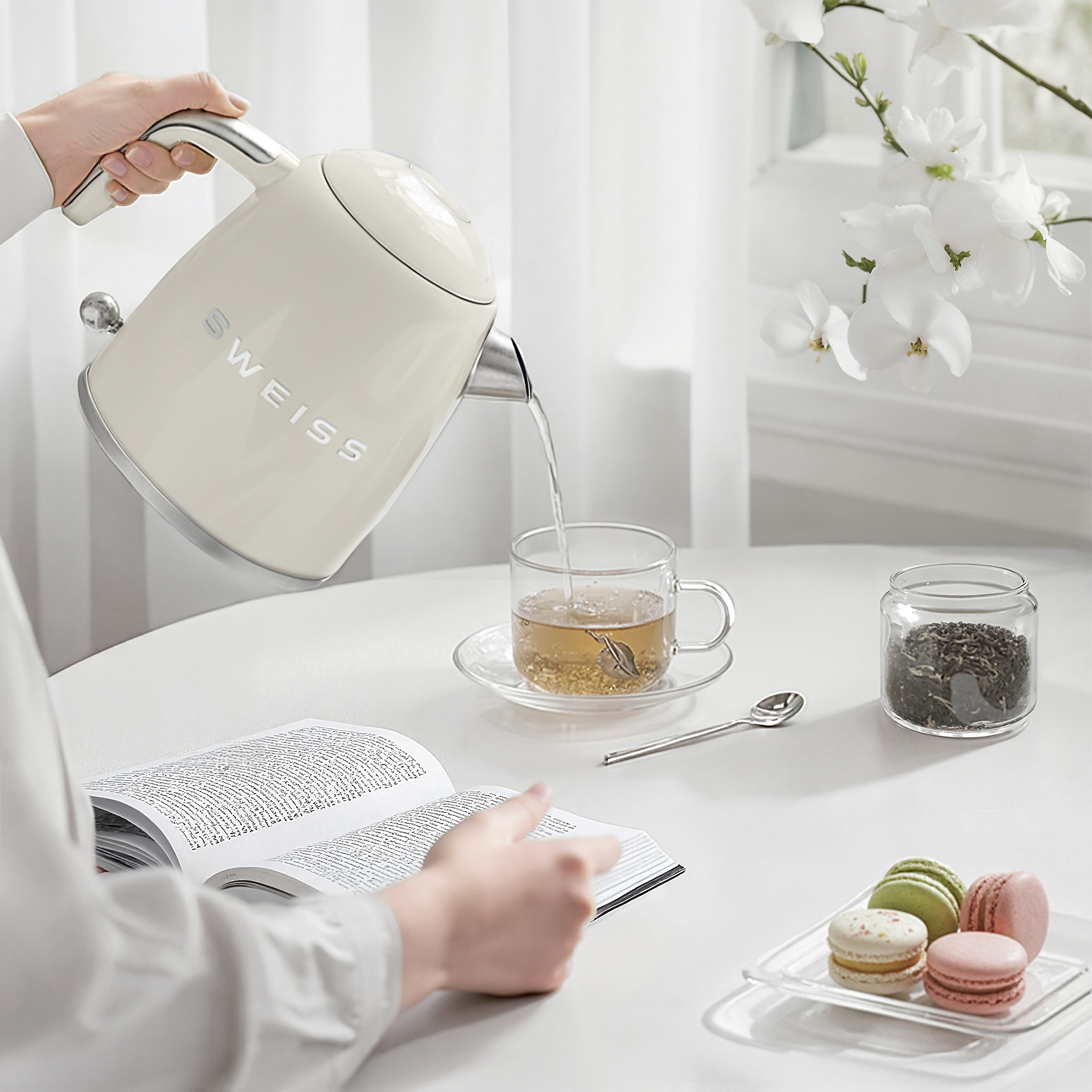 Person gießt Wasser aus einem beigen Wasserkocher in eine Teetasse auf einem Tisch mit Buch und Makronen.