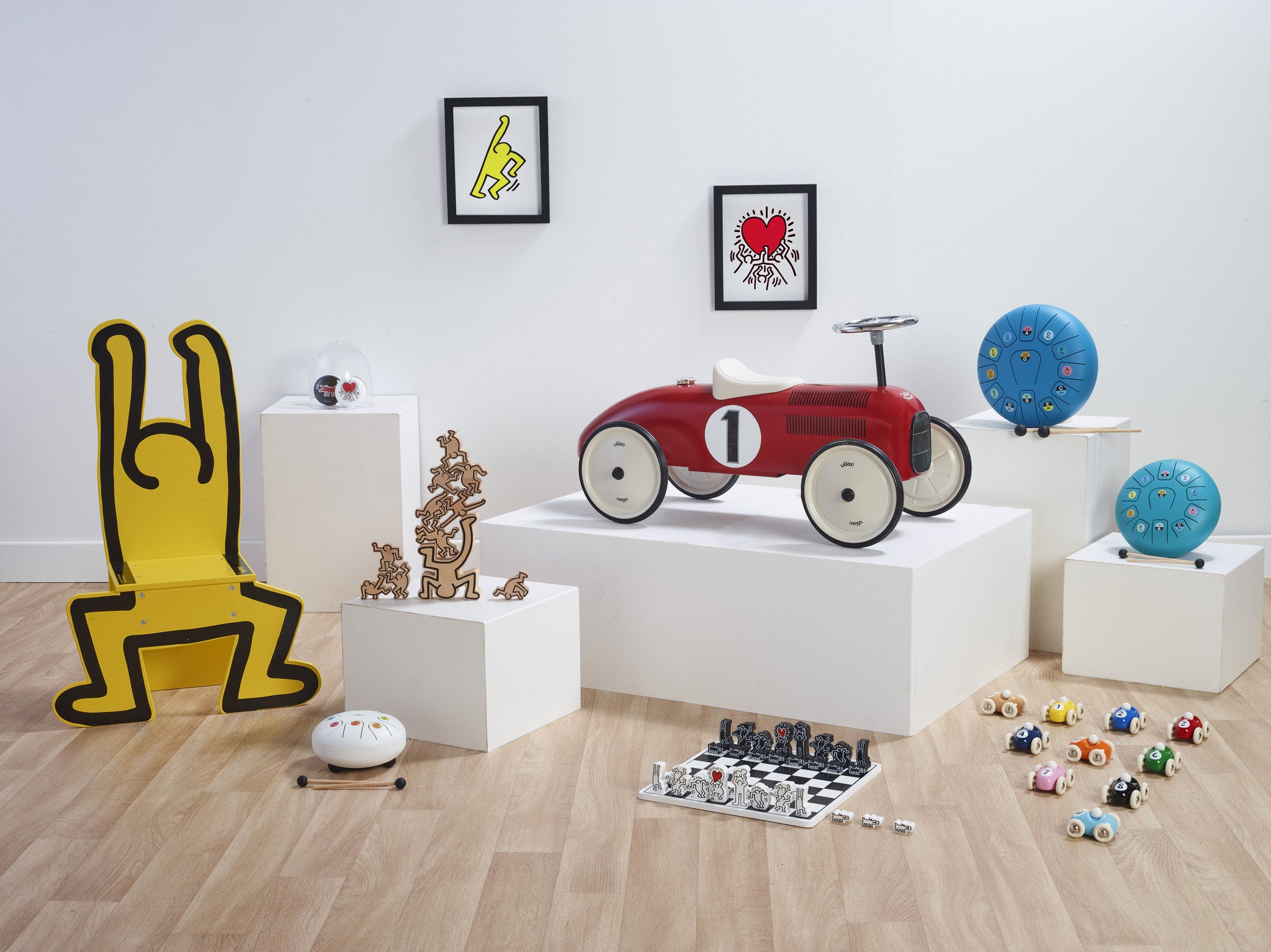 Ein rotes Spielzeugauto, eine gelbe Stuhlfigur und verschiedene Holzspielzeuge auf weißen Podesten.