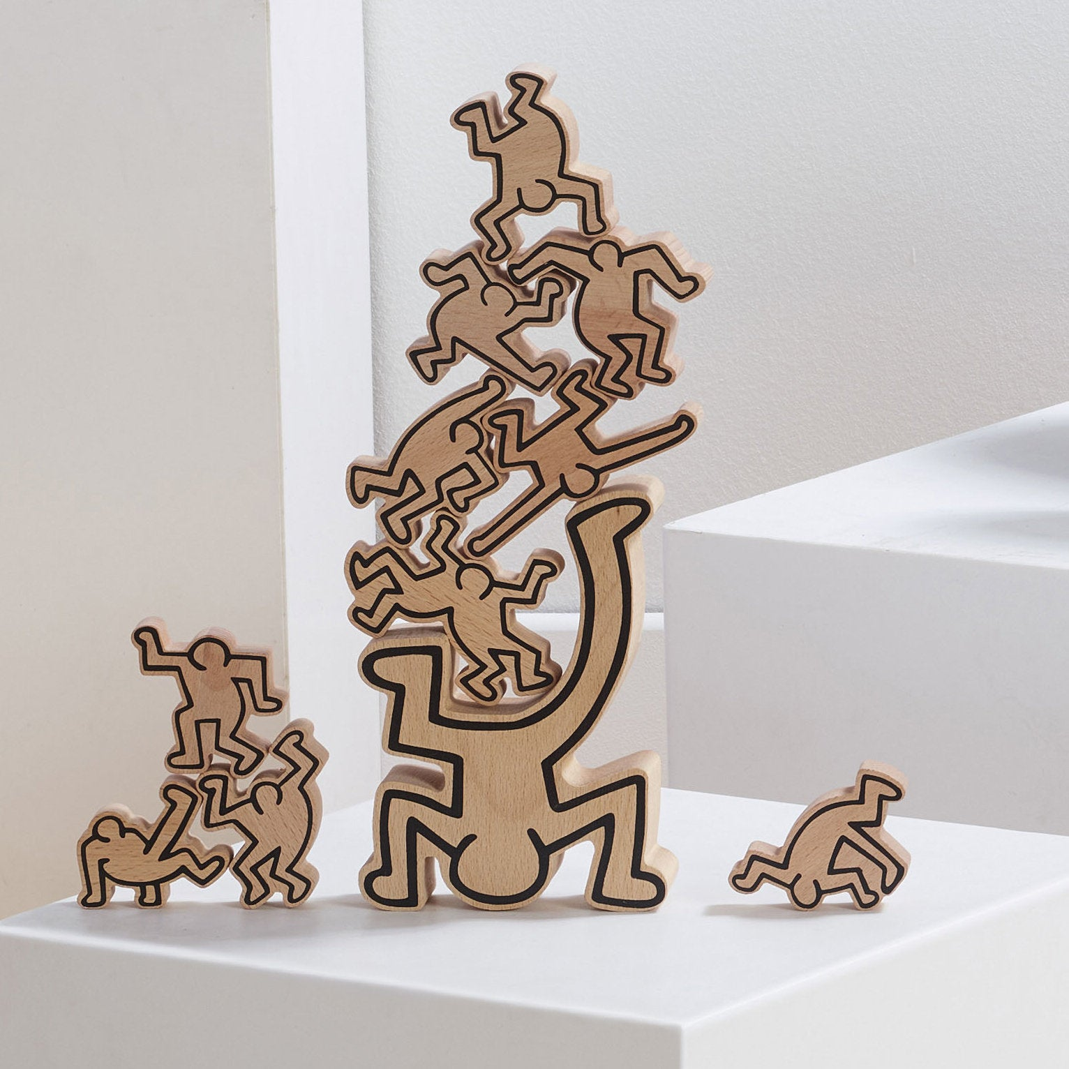 Holzfiguren im Keith-Haring-Stil, gestapelt auf einer weißen Oberfläche vor hellem Hintergrund.