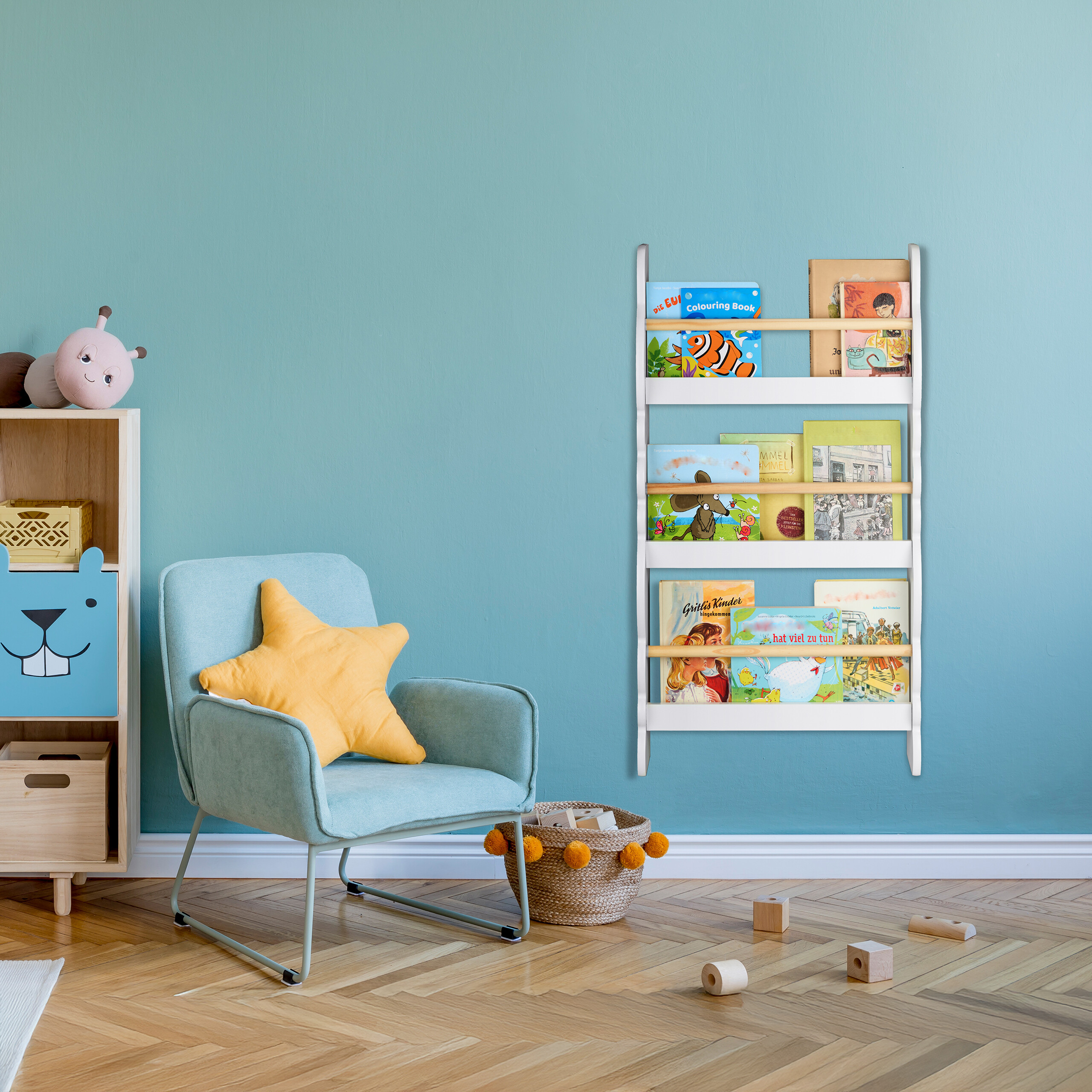 Ein Kinderzimmer mit blauem Sessel, gelbem Kissen, Wandregal voller Bücher und Holzspielzeug auf Parkettboden.