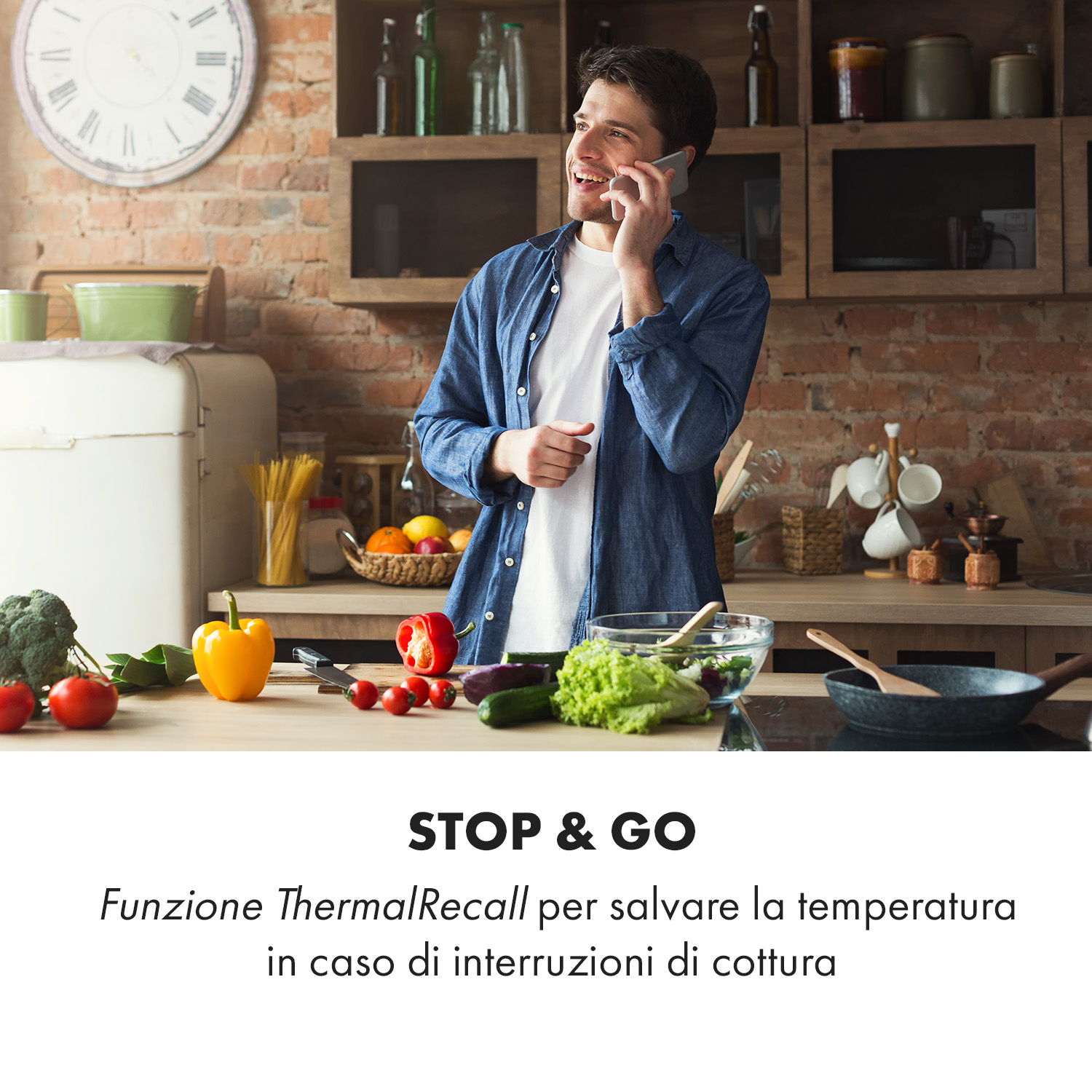 Un uomo con una camicia blu parla al telefono in una cucina con verdure sul bancone e una parete in mattoni.