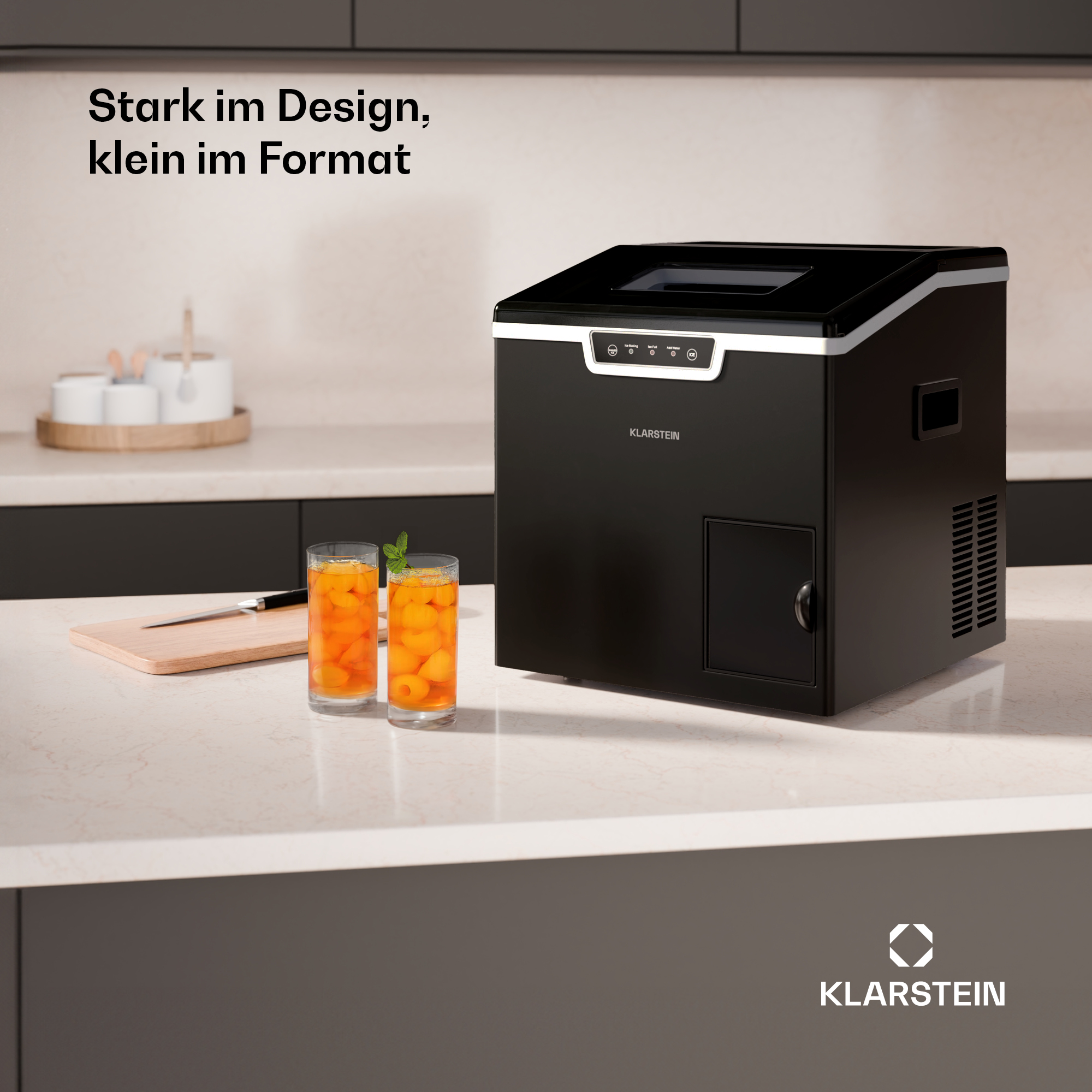 Schwarze Eiswürfelmaschine auf einer Küchenarbeitsplatte neben zwei Gläsern mit orangem Getränk und Minze.
