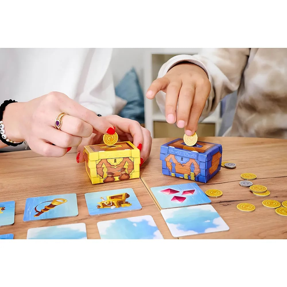 Zwei Personen spielen ein Brettspiel, stecken Goldmünzen in kleine Schatztruhen und ordnen Karten auf dem Holztisch an.