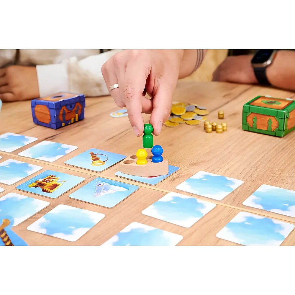 Person setzt grüne Spielfigur auf ein Holzboot mit gelben und blauen Figuren, umgeben von Spielkarten auf einem Tisch.