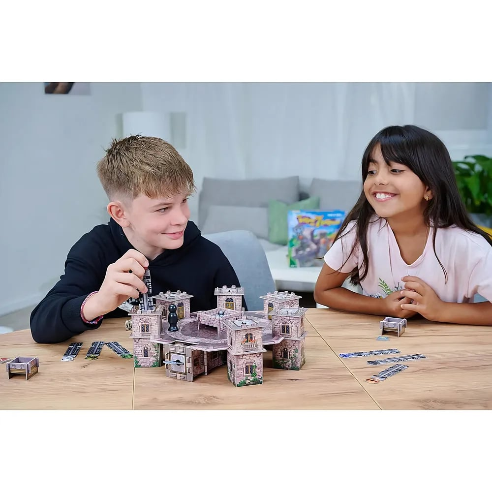Zwei Kinder lächeln, während sie an einem Holztisch in einem Wohnzimmer mit einem Pappburg-Brettspiel spielen.