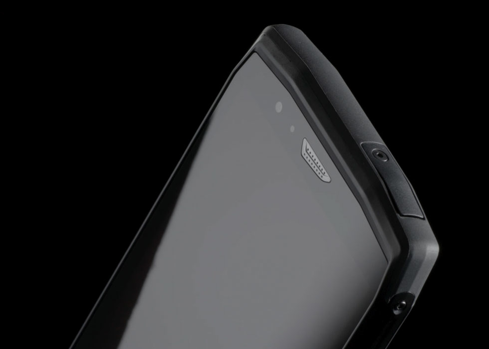 Primer plano de la esquina superior de un teléfono resistente, con funda negra, pantalla, auricular y cámara sobre fondo negro.