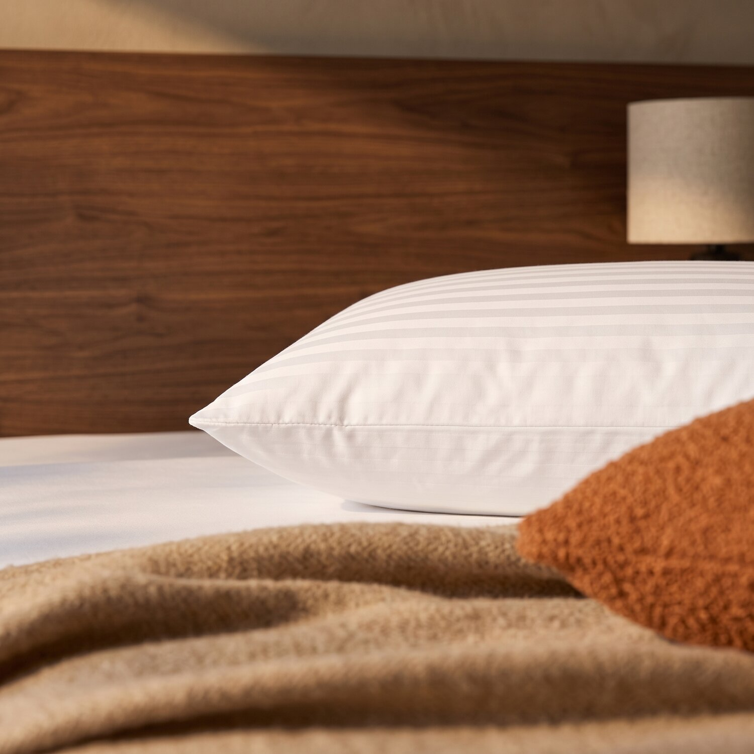 Una almohada blanca a rayas en una cama con cabecera de madera marrón, una manta acogedora y una lámpara pequeña.