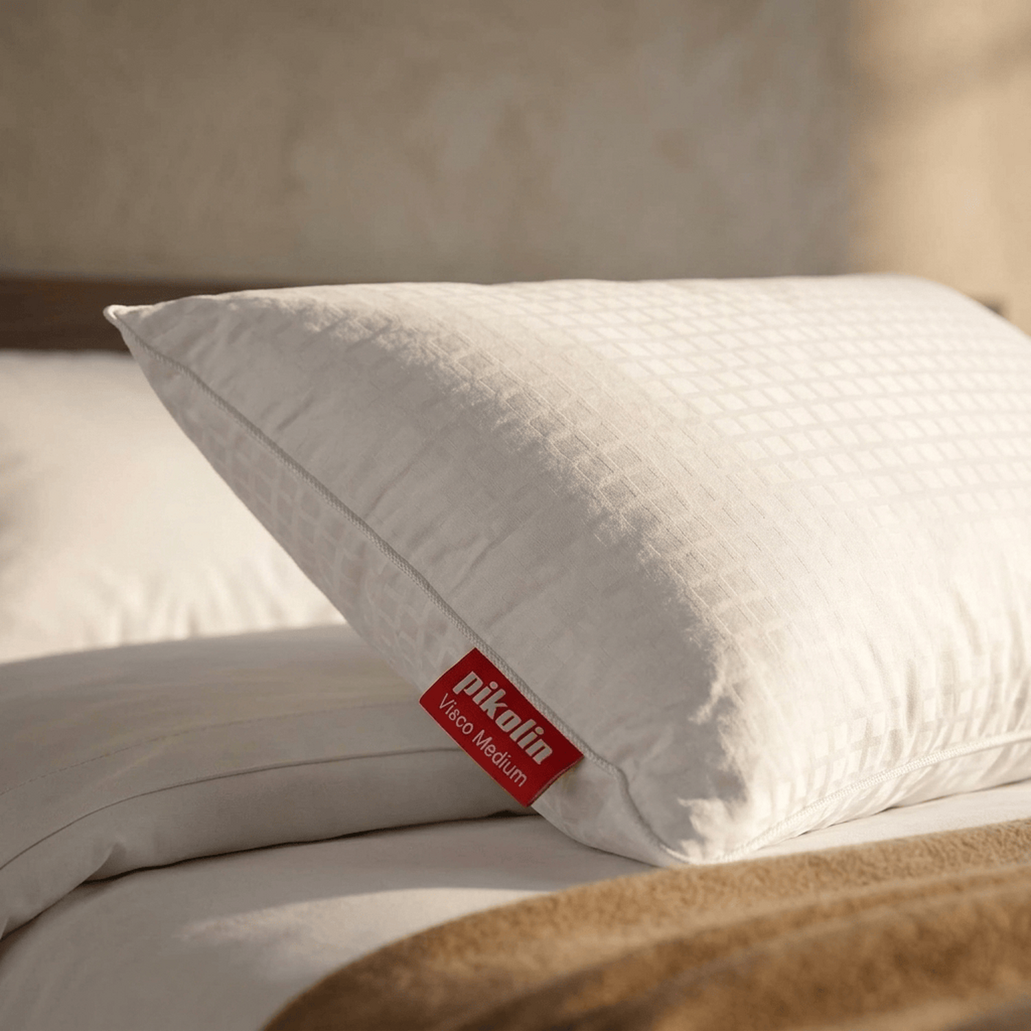 Una almohada blanca Pikolin Visco Medium descansa en una cama con textiles beiges.