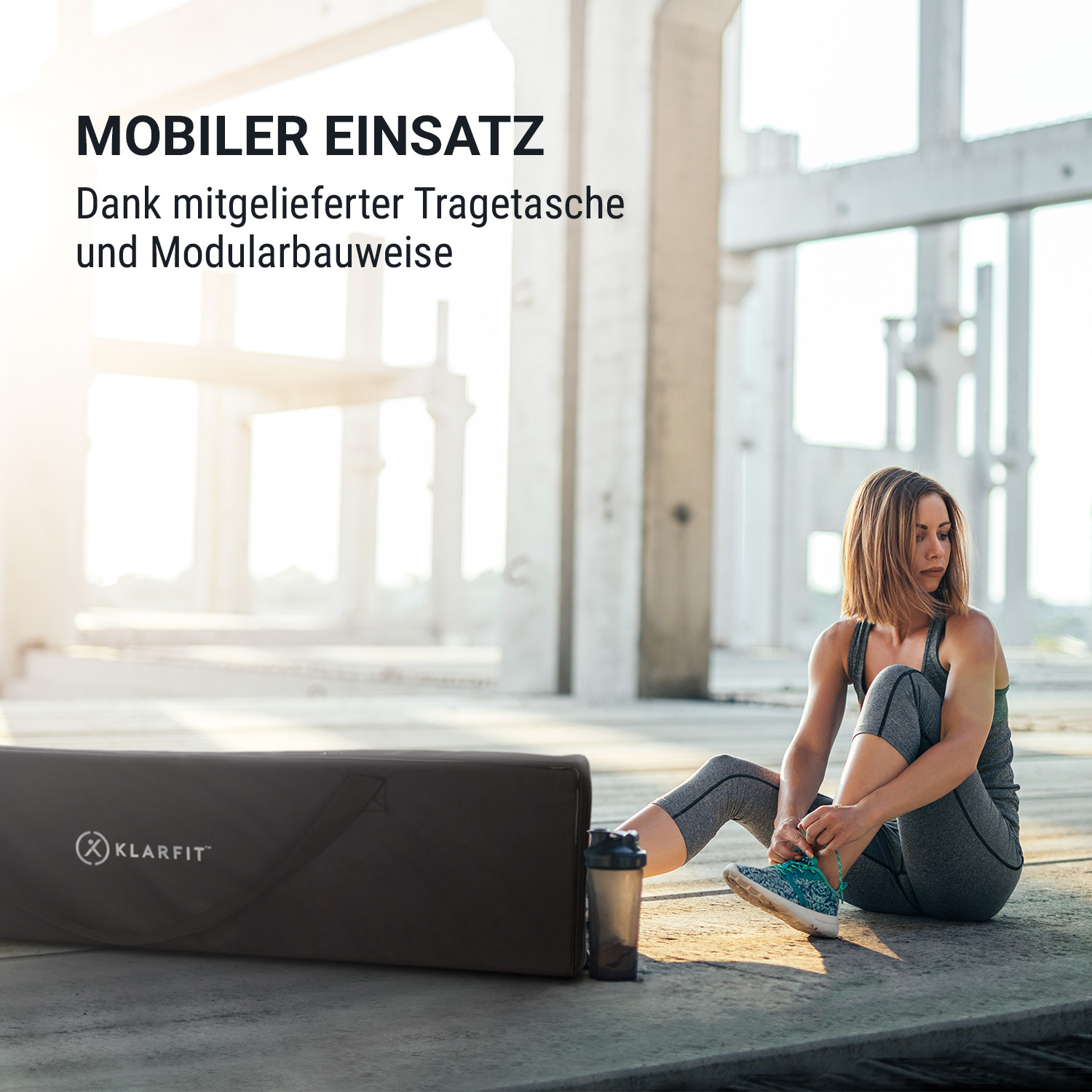 Eine Frau sitzt in einer Industriehalle auf dem Boden und bindet ihre Sportschuhe neben einer grauen Tasche.