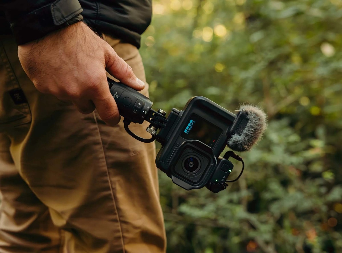 Una persona sostiene una cámara GoPro negra en un palo selfie al aire libre con follaje verde.