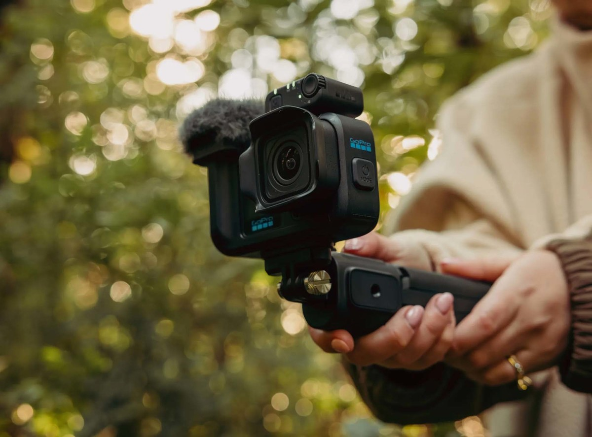 Una persona sostiene una cámara GoPro negra con micrófono y mango al aire libre con fondo verde.