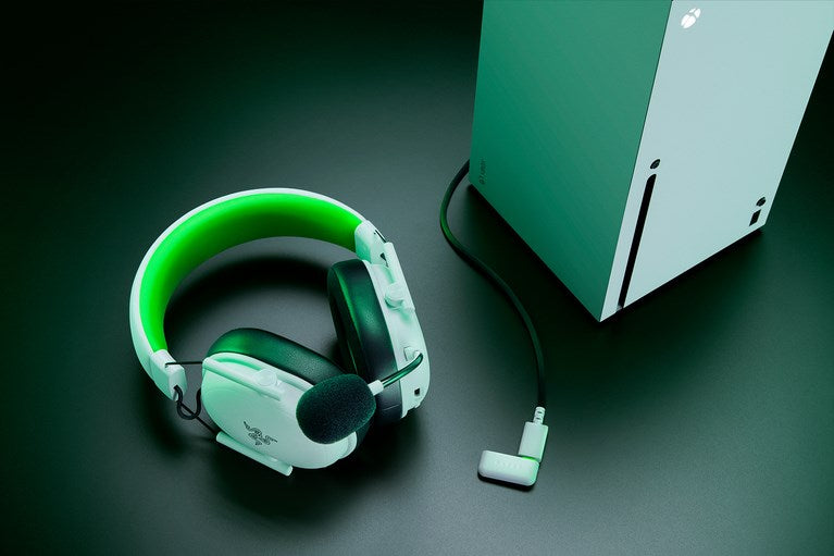 Auriculares de juego blancos y verdes con micrófono y cable negro junto a una Xbox Series X sobre fondo verde.