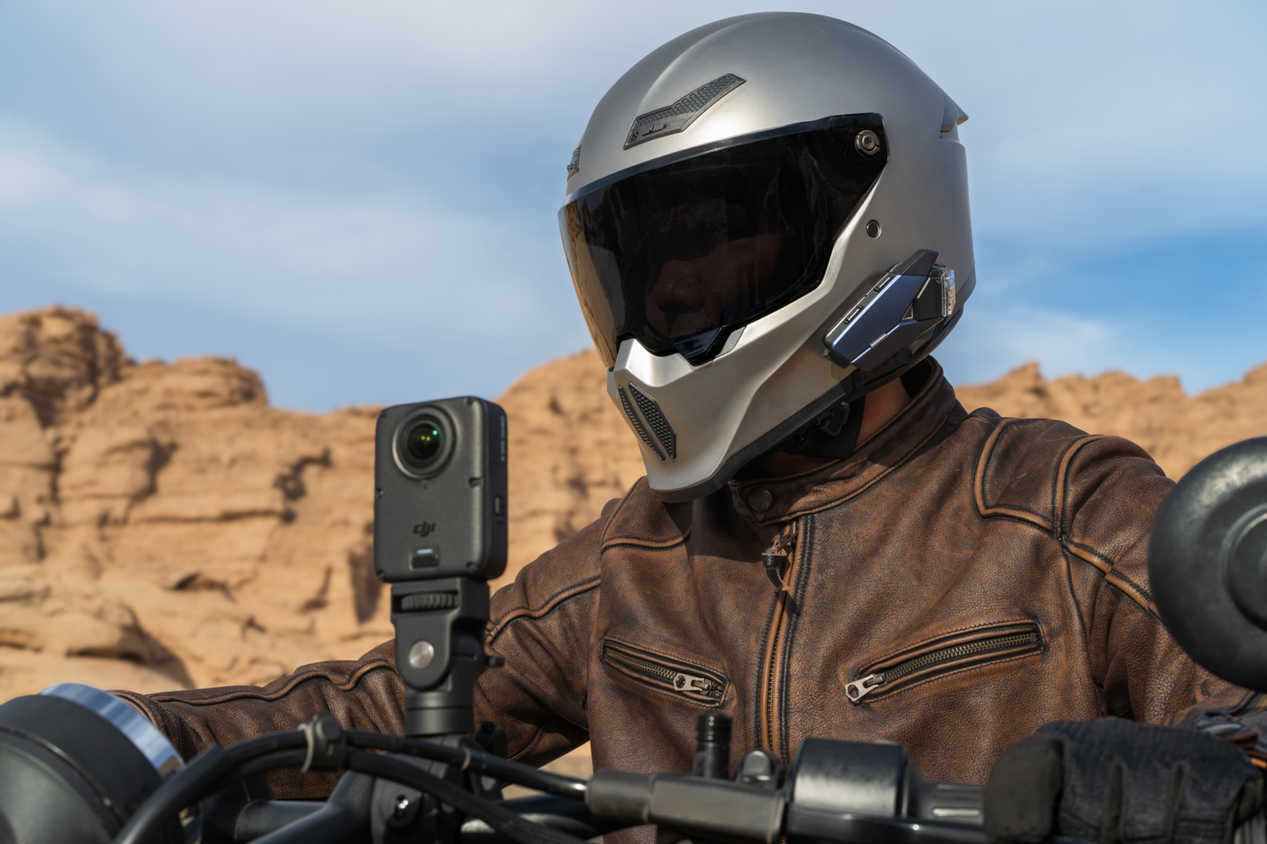 Motociclista con casco plateado y chaqueta de cuero marrón frente a paisaje rocoso con cámara montada.