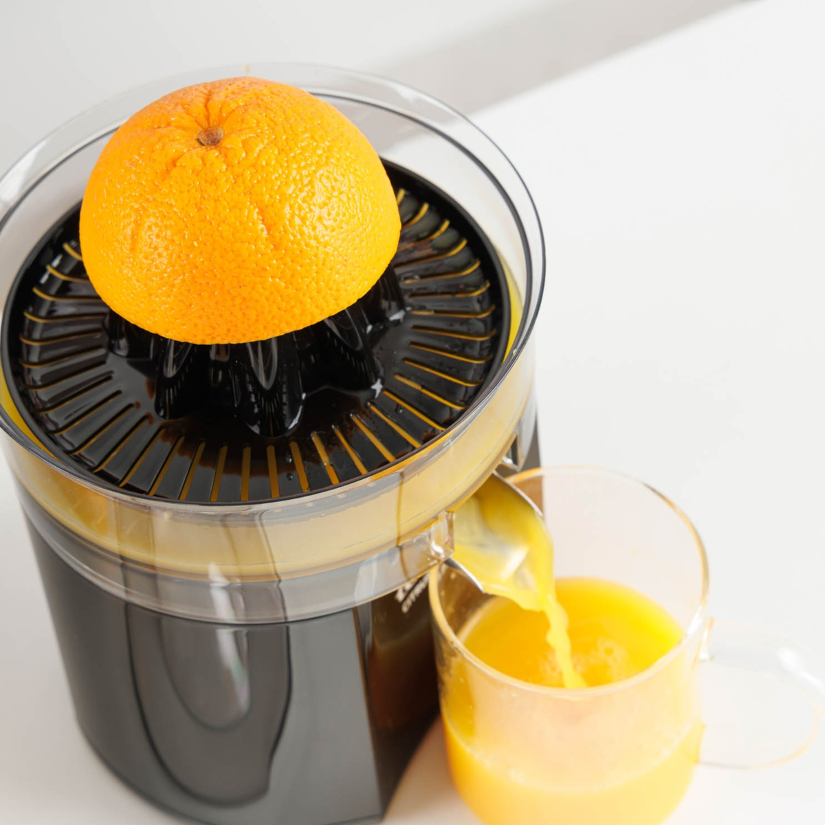 Un exprimidor exprime media naranja, el jugo fresco fluye hacia un vaso en una superficie blanca.