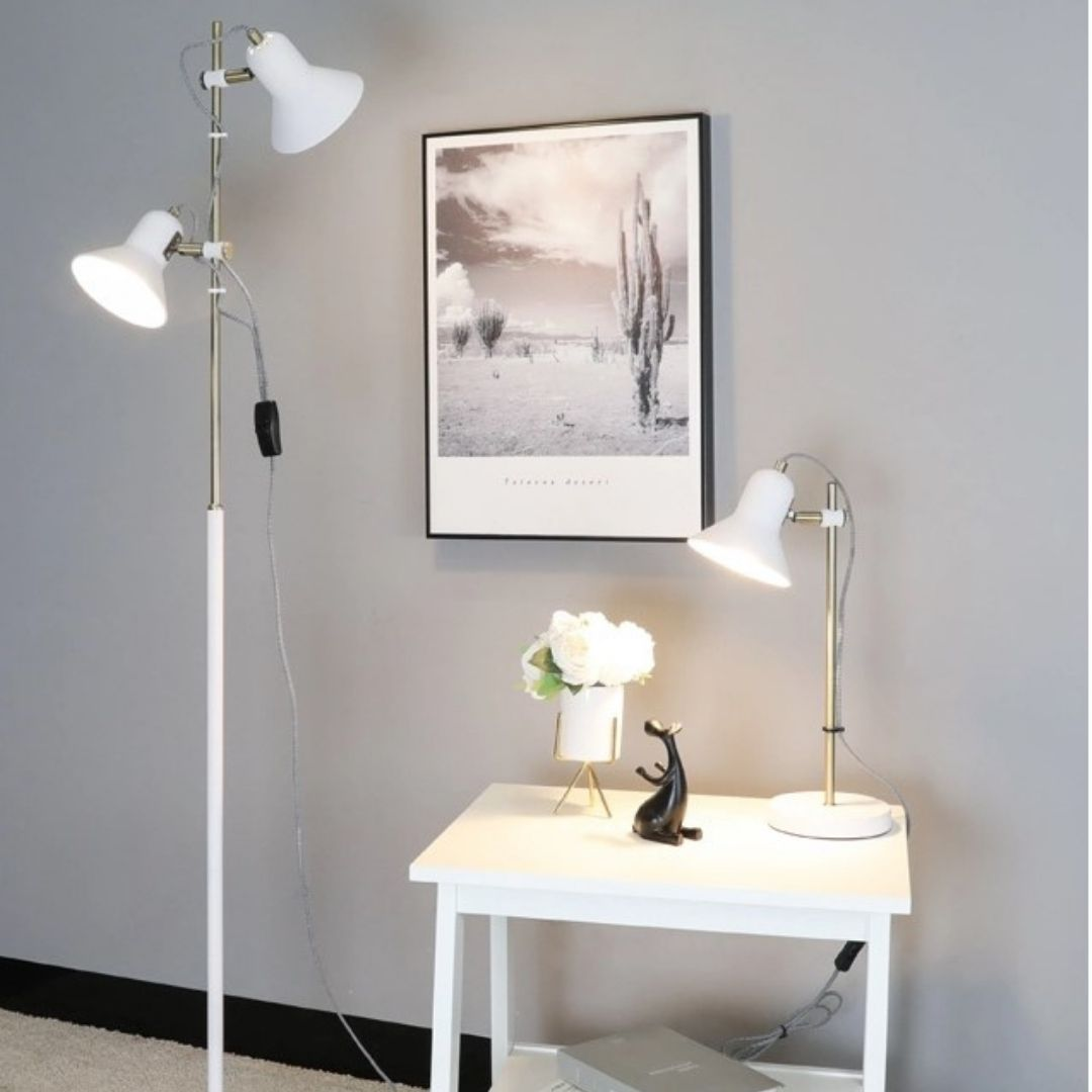 Una habitación con mesa auxiliar blanca, lámpara de mesa encendida, lámpara de pie, jarrón con flores y foto del desierto.