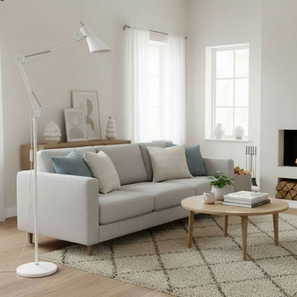 Una sala luminosa con sofá gris, lámpara de pie blanca, mesa de centro de madera, alfombra y ventanas con cortinas.