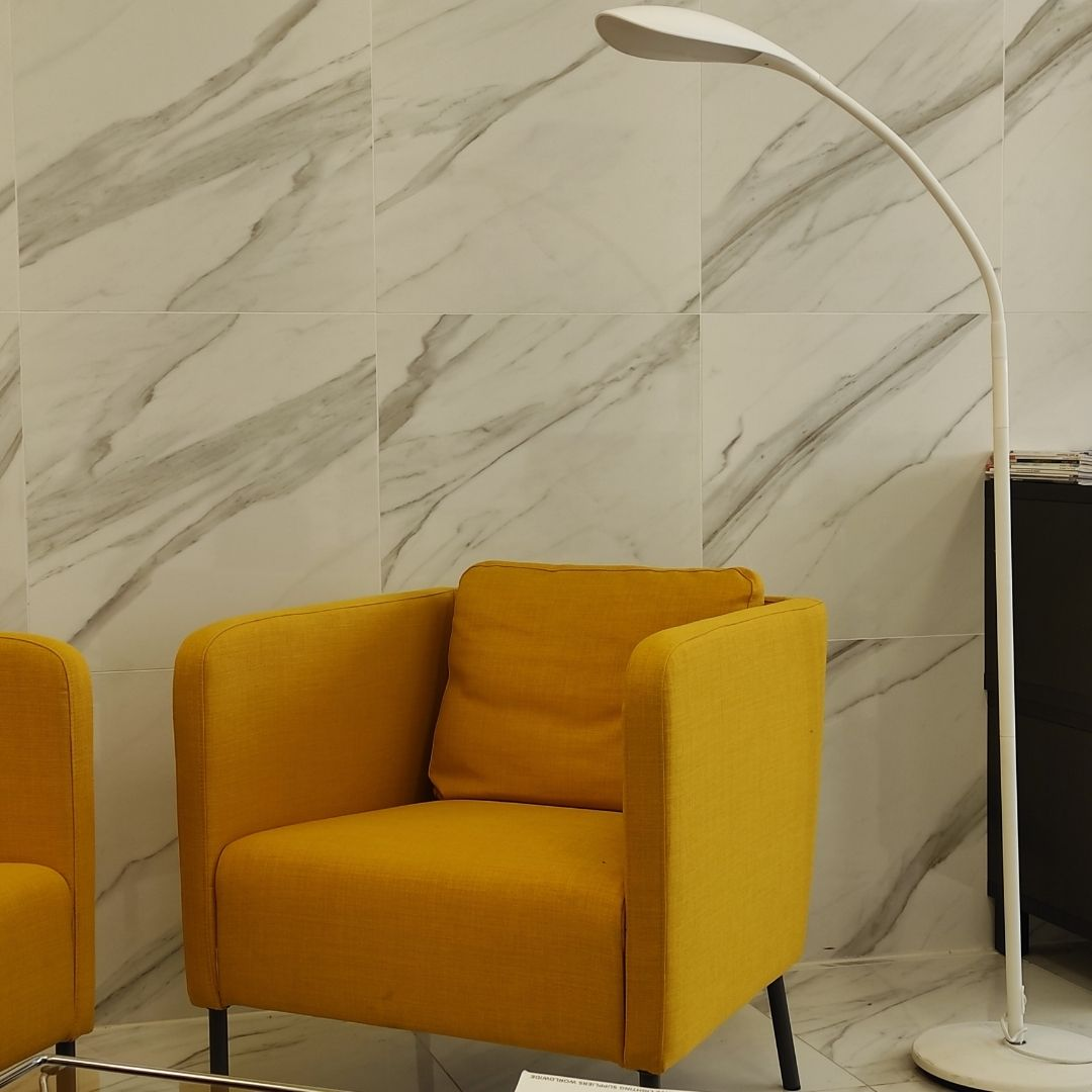 Un sillón amarillo frente a una pared de mármol blanco y gris, con una lámpara de pie arqueada blanca.