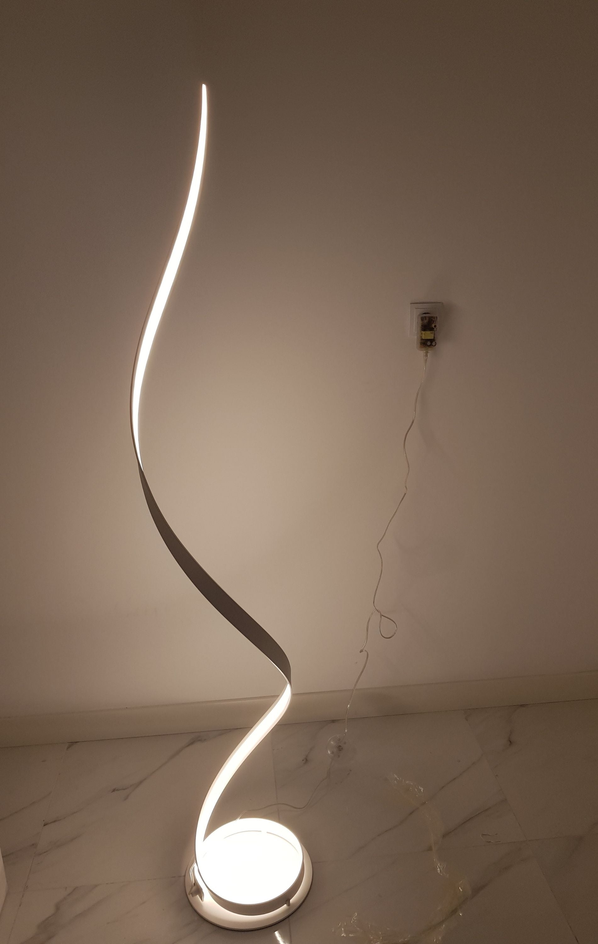 Una lámpara de pie moderna y curvada brilla en un suelo de mármol contra una pared blanca.