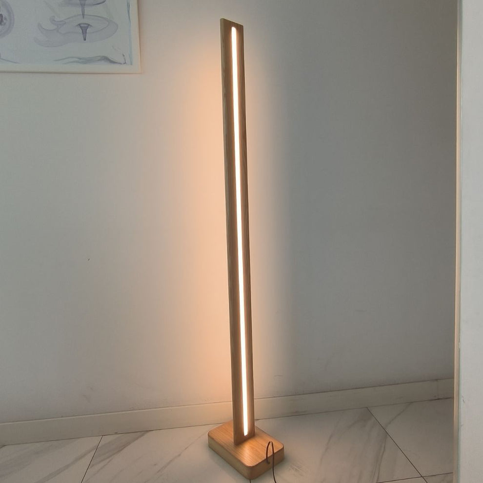 Una lámpara de pie de madera con una franja de luz brillante sobre un suelo de baldosas claras junto a una pared blanca.