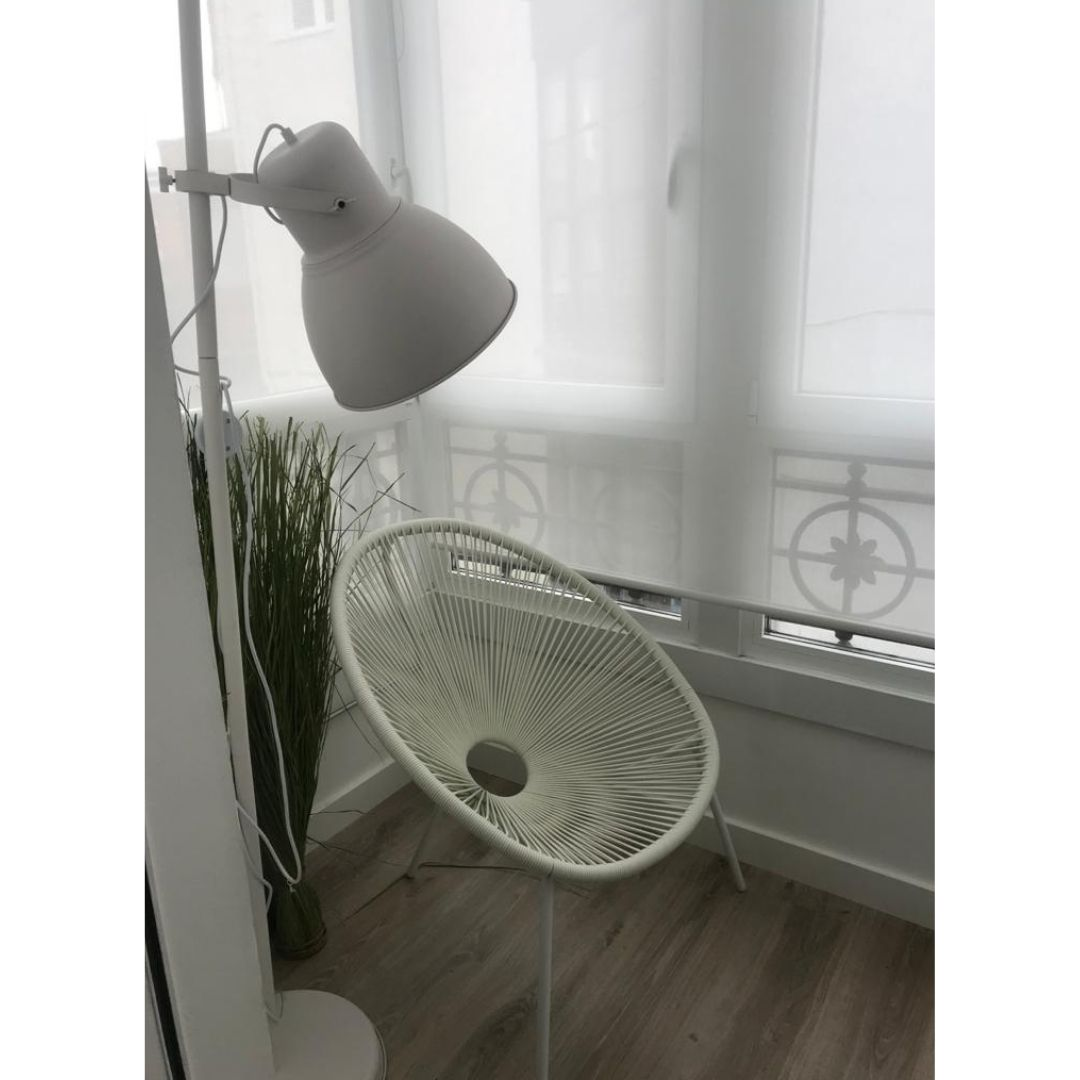 Una silla blanca de cuerdas junto a una ventana luminosa, con una lámpara de pie y una planta alta.