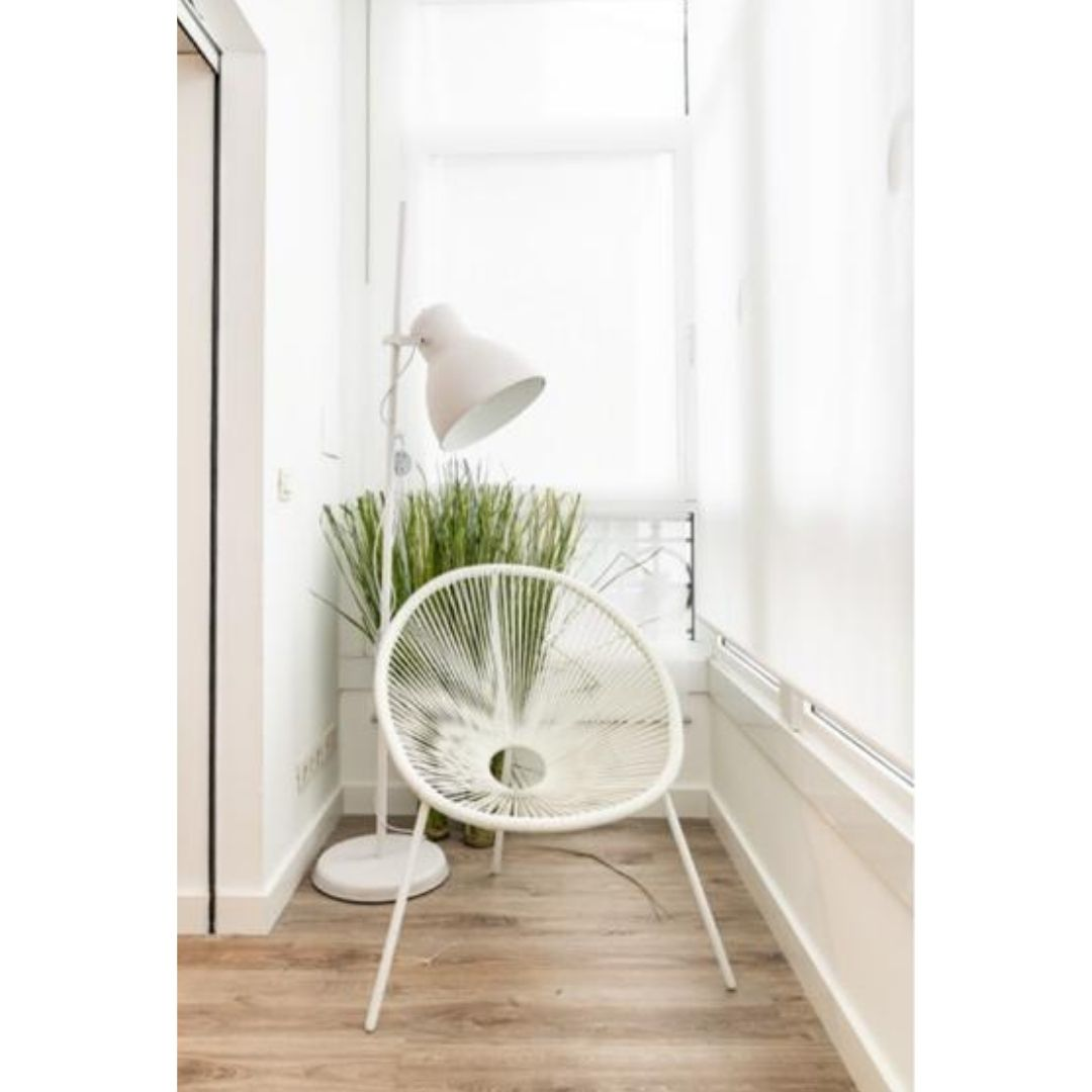 Una silla blanca, una lámpara de pie y plantas verdes se encuentran en una habitación luminosa frente a una gran ventana.