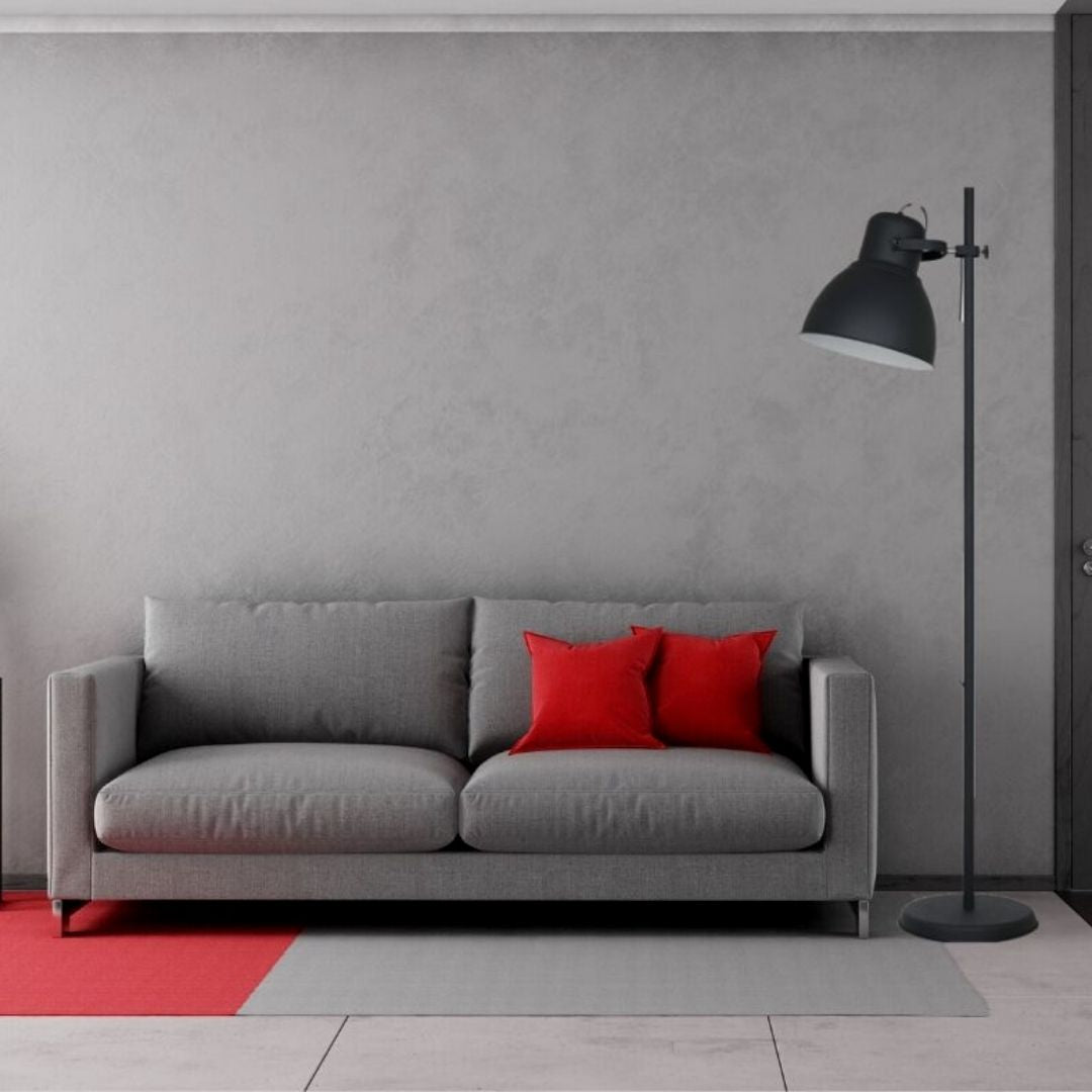 Un sofá gris con dos cojines rojos, una lámpara de pie negra y una alfombra roja contra una pared gris.