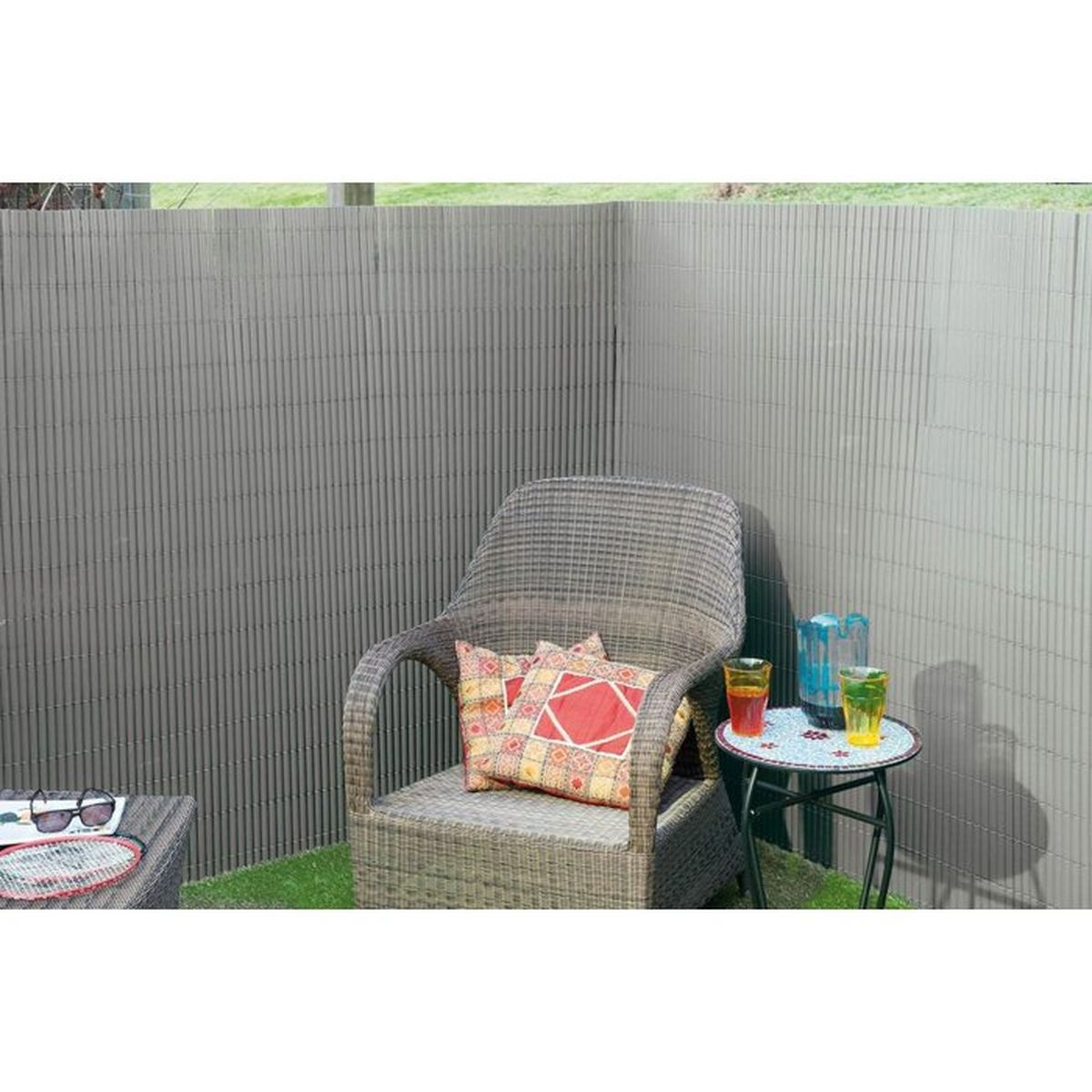 Grauer Rattansessel mit bunten Kissen und einem kleinen Tisch vor einem Sichtschutzzaun.