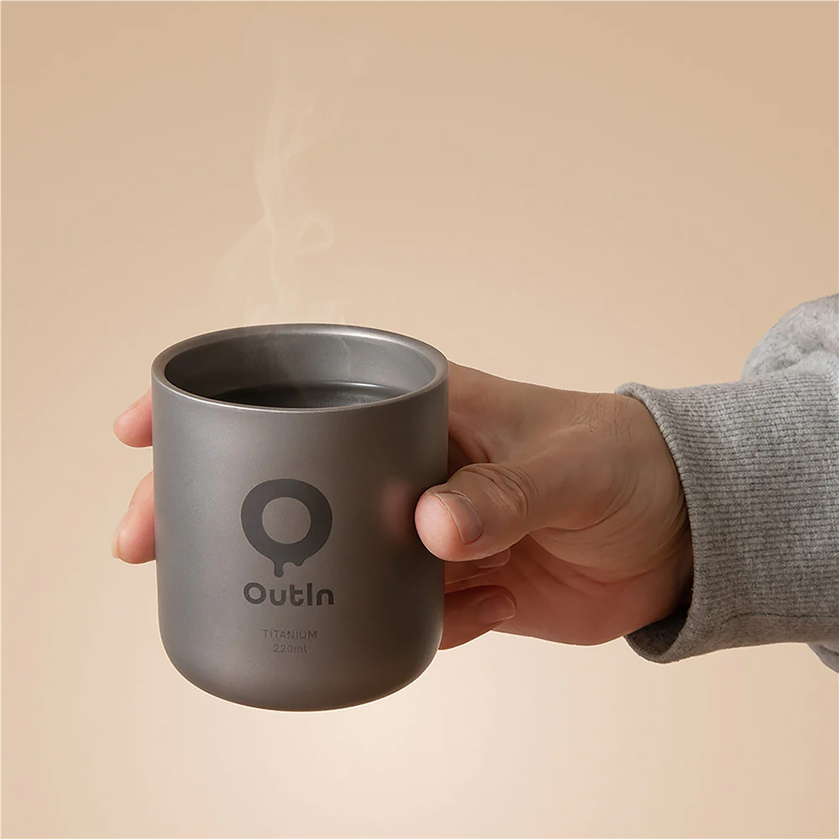 Una mano sostiene una taza de titanio Outin gris con vapor ascendente sobre fondo bege.