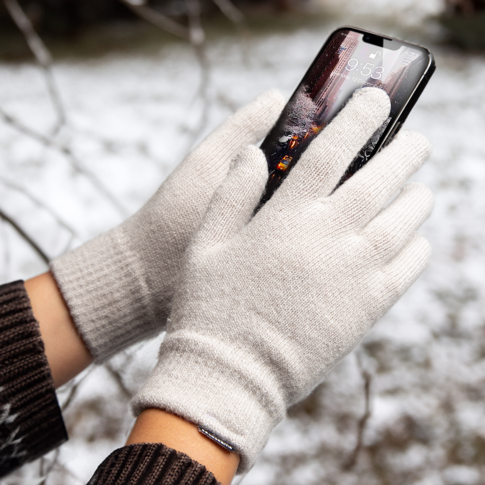 Manos con guantes de punto grises sostienen un smartphone sobre fondo nevado.