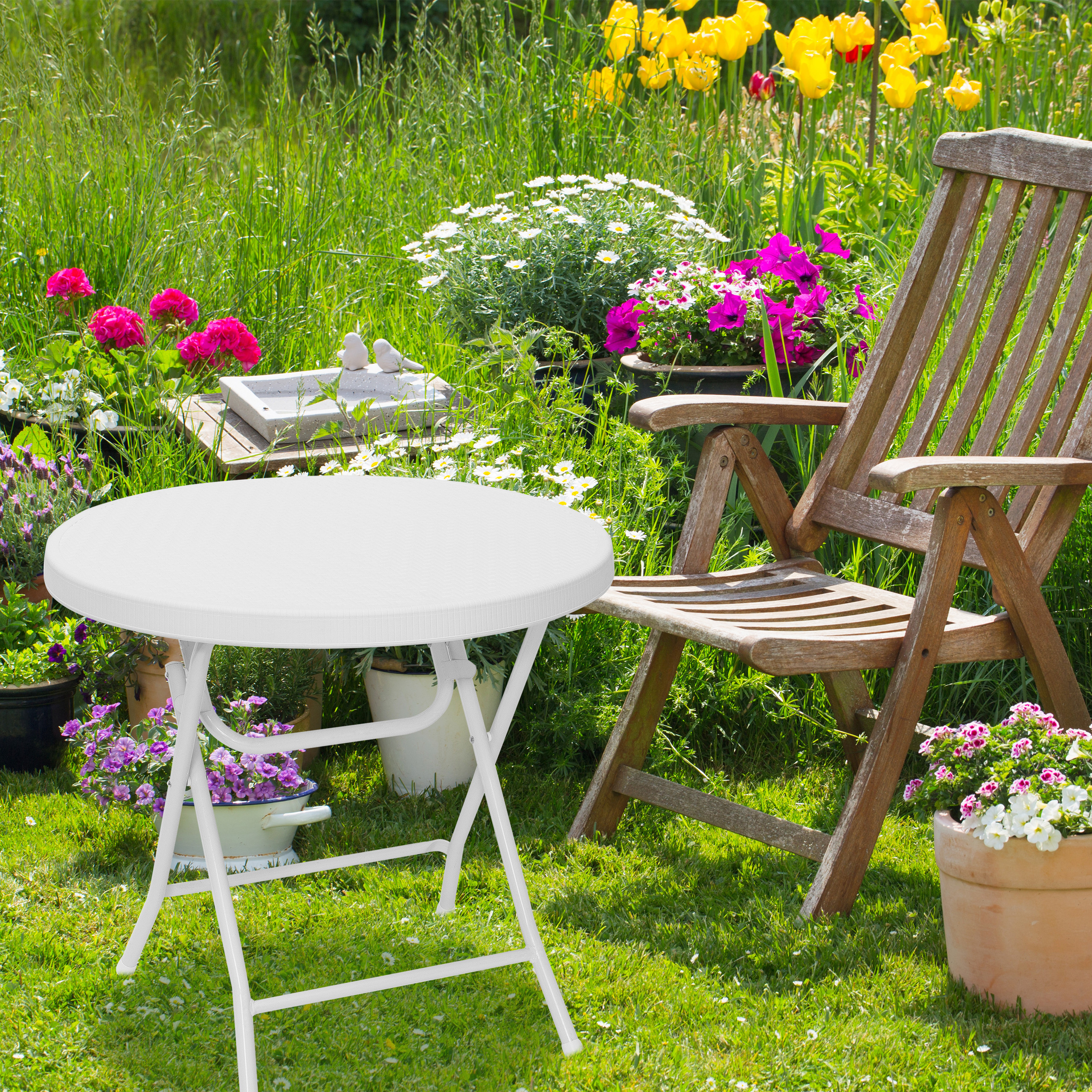 Ein weißer Klapptisch und ein Holzstuhl stehen auf einer grünen Wiese, umgeben von bunten Blumen und üppigem Grün.