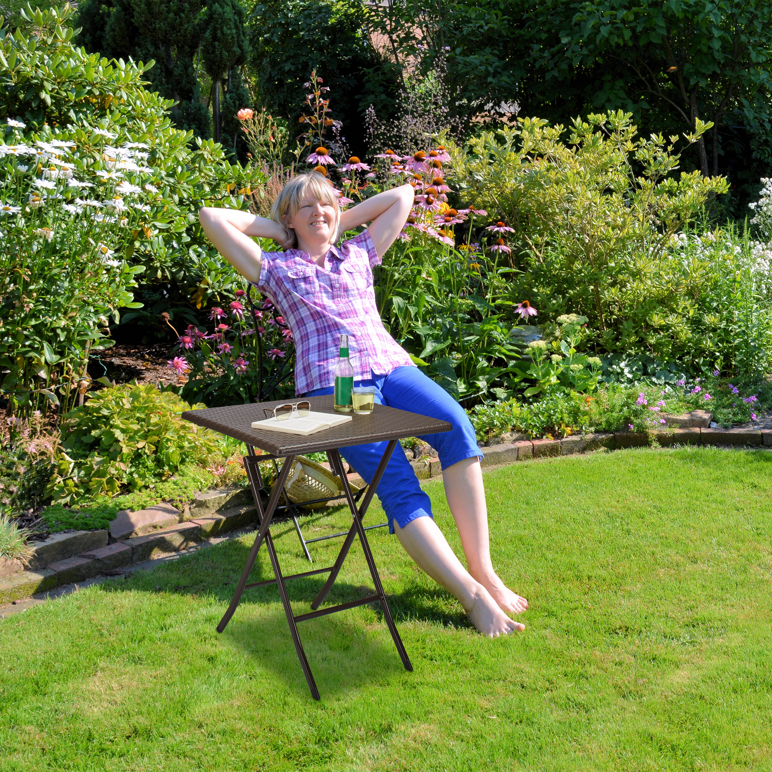 Frau entspannt in einem sonnigen Garten mit Blumen im Hintergrund und einem kleinen Tisch im Vordergrund.