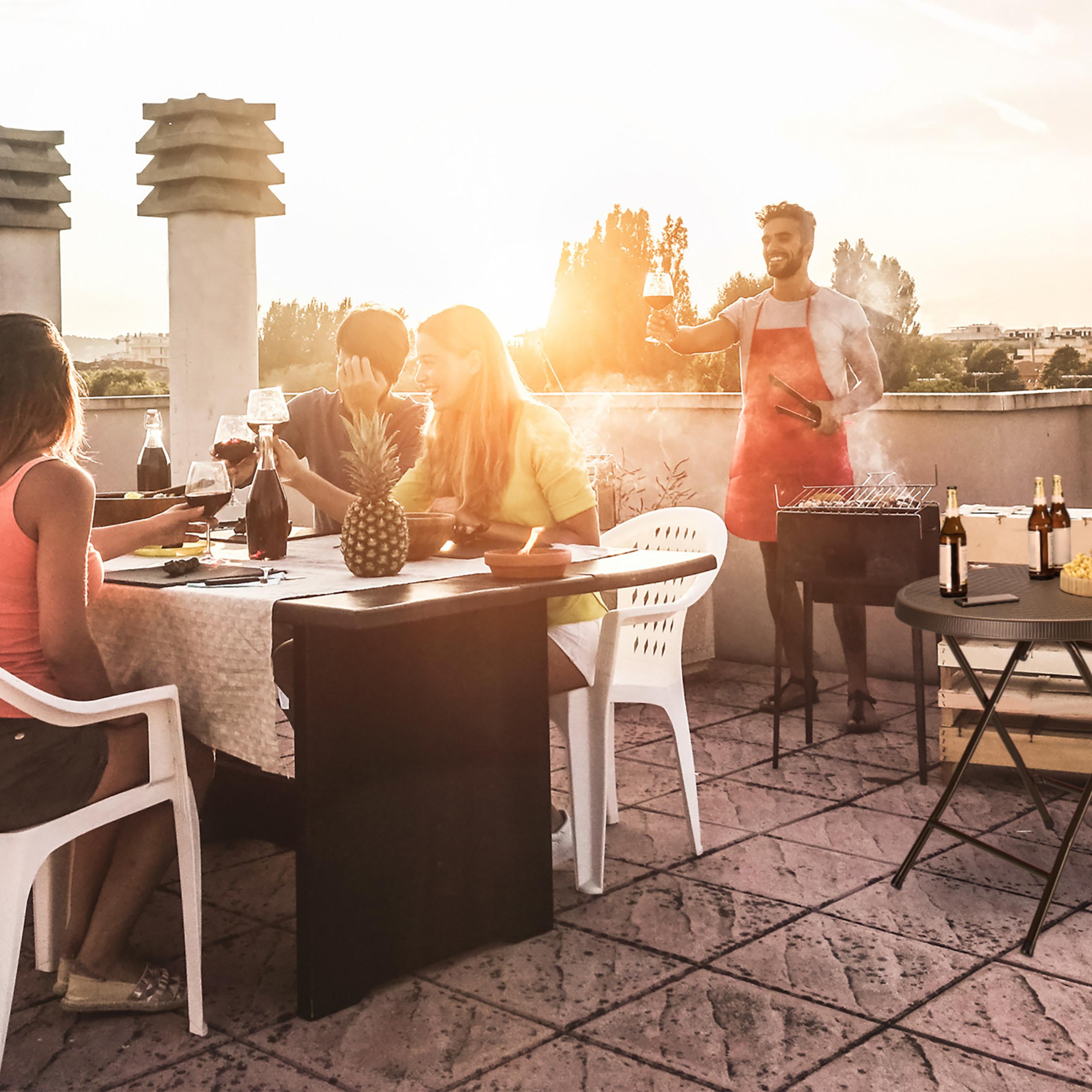 Freunde genießen eine Dachterrasse-Grillparty bei Sonnenuntergang, mit einem grillenden Mann.