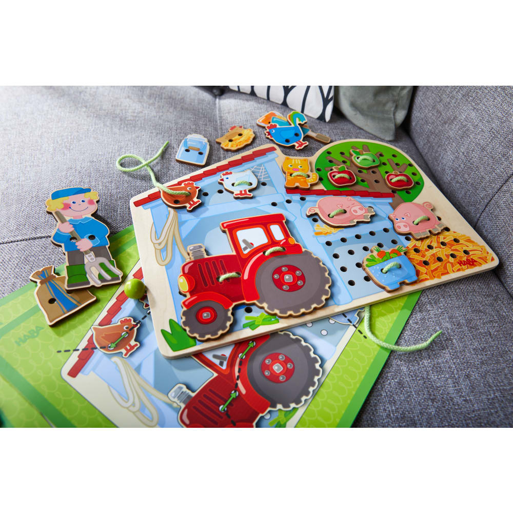 Holz-Schnürspiel mit Bauernhof-Motiven, rotem Traktor und Tieren auf einer grauen Couch.