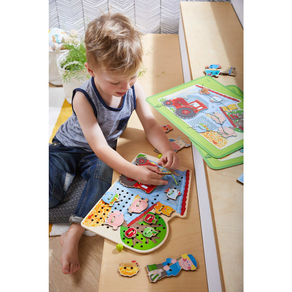 Ein Junge sitzt auf dem Boden und spielt mit einem bunten Holz-Fädelspiel mit Bauernhof-Motiven.