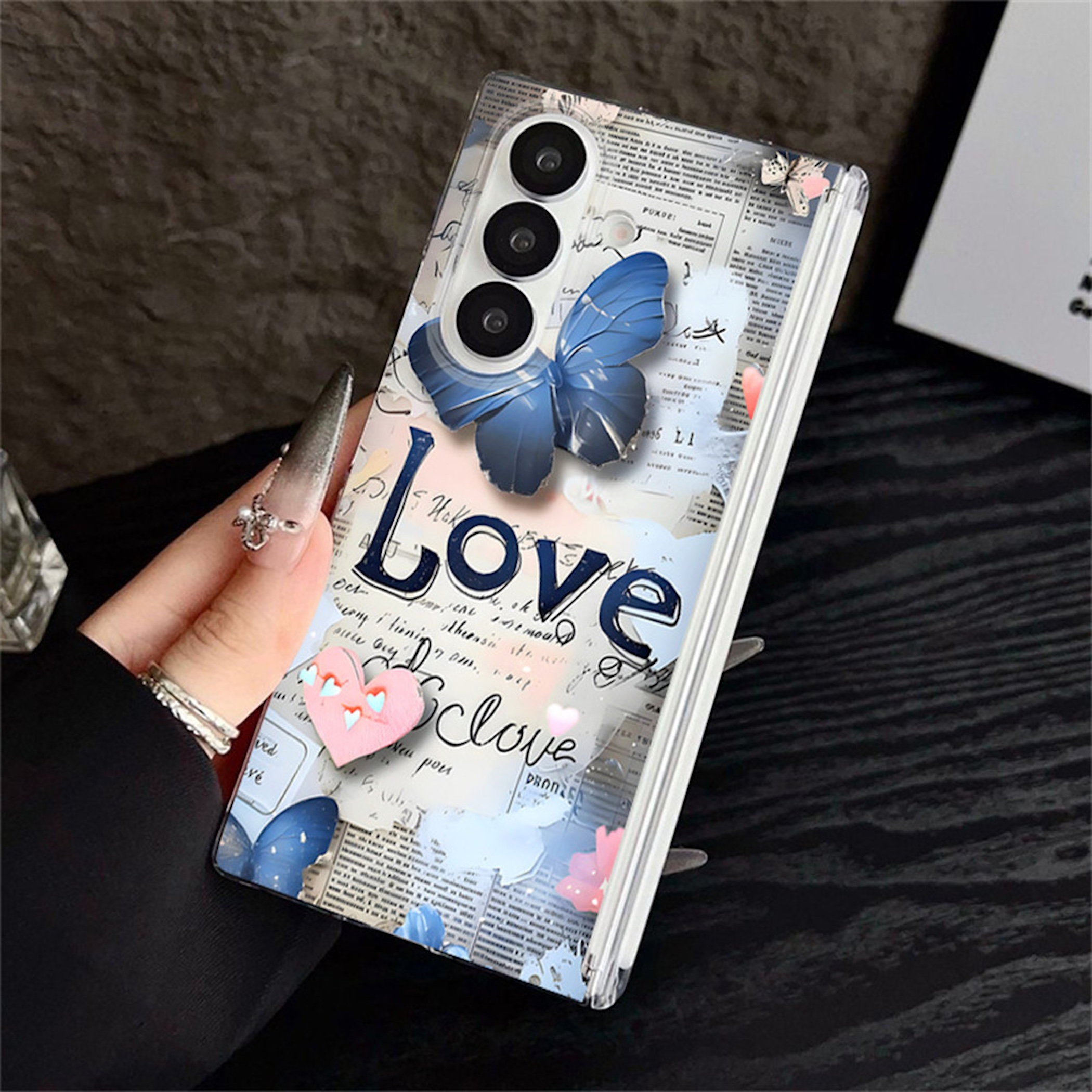 Eine Hand hält ein Handy mit dekorativer Hülle mit blauen Schmetterlingen, Love-Schriftzug und Zeitungsmotiv.