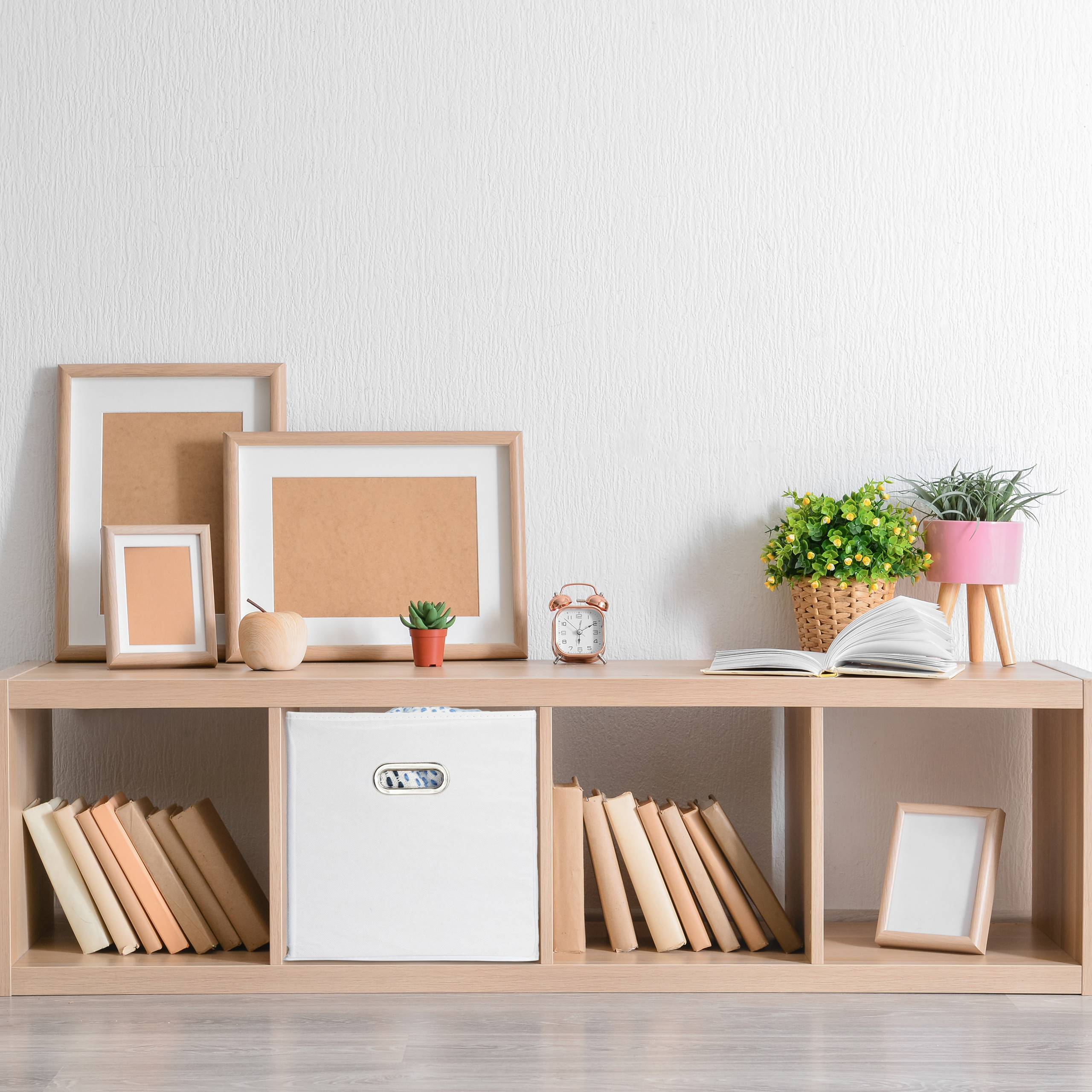 Ein hölzernes Regal mit Bilderrahmen, Büchern, Pflanzen, einem Wecker und einer Aufbewahrungsbox vor weißer Wand.