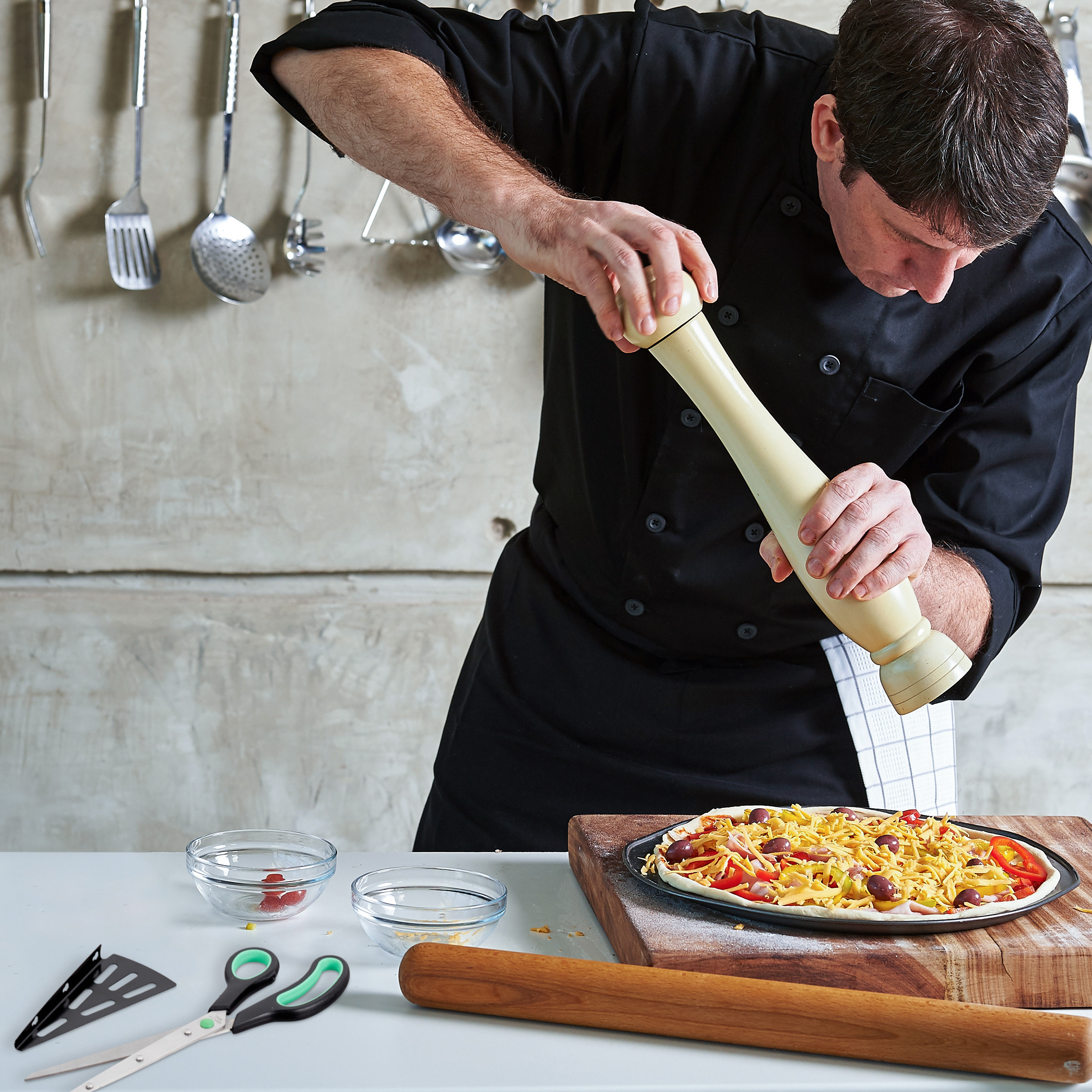 Ein Koch in schwarzer Uniform mahlt Gewürze über eine ungebackene Pizza in einer Küche.